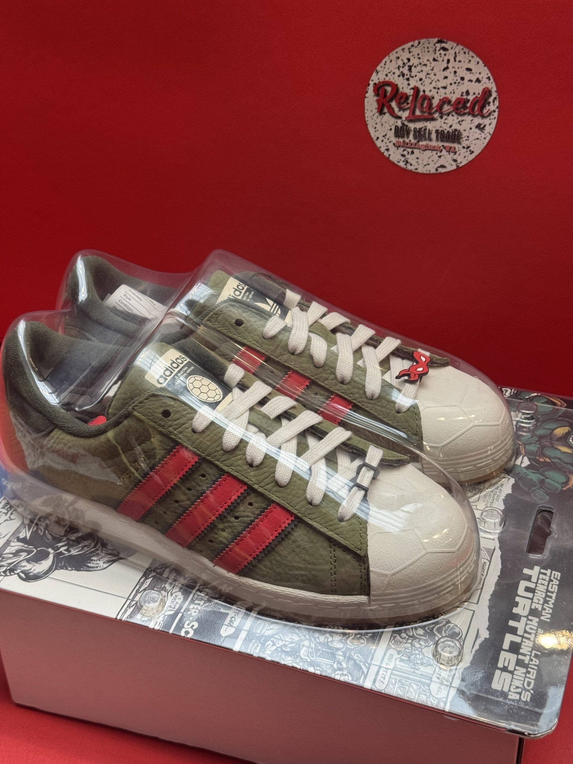 Pair of olive green Adidas Superstar sneakers with red stripes, in plastic, on a box against a red background.