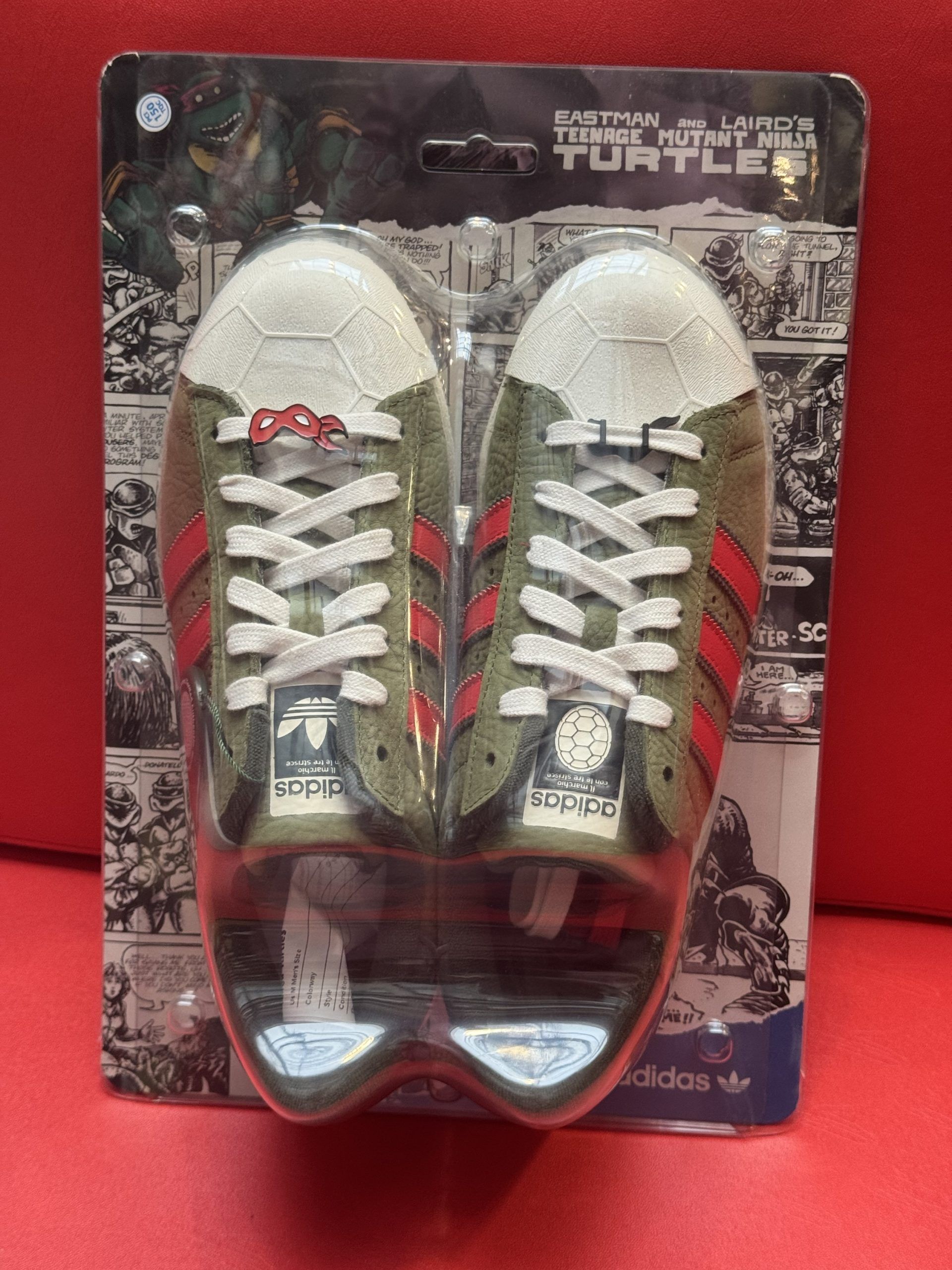 Pair of olive green Adidas Superstar sneakers with red stripes, in plastic, on a box against a red background.