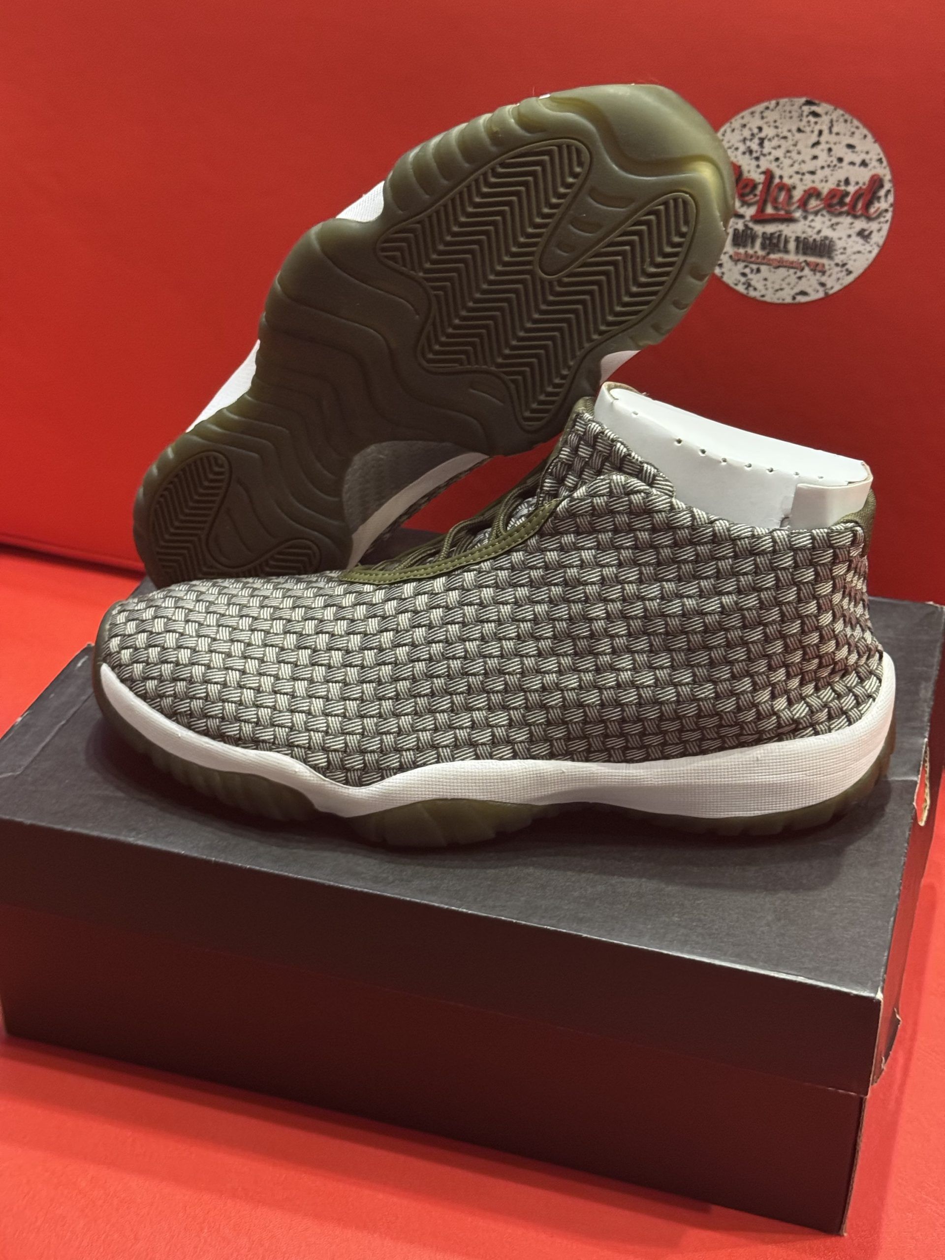Pair of woven-pattern sneakers, olive green sole and trim, sitting on a black shoebox, against a red background.