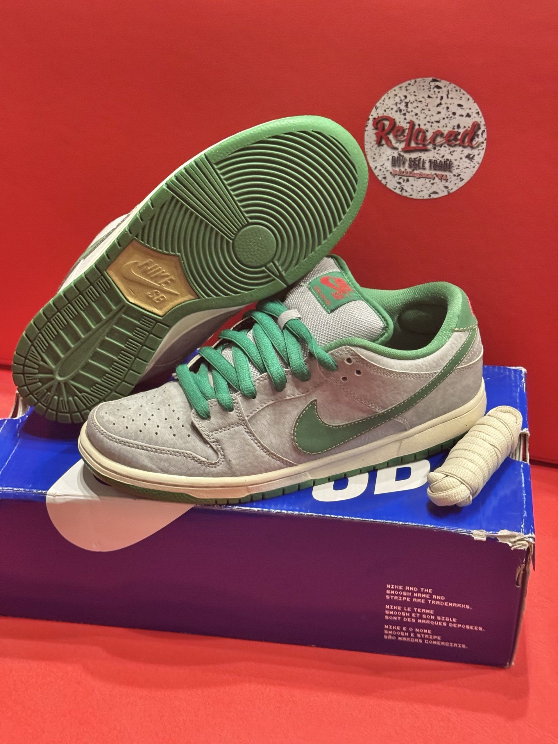 Nike SB Dunk Low sneakers in gray, green, and cream, displayed with the box and a red background.