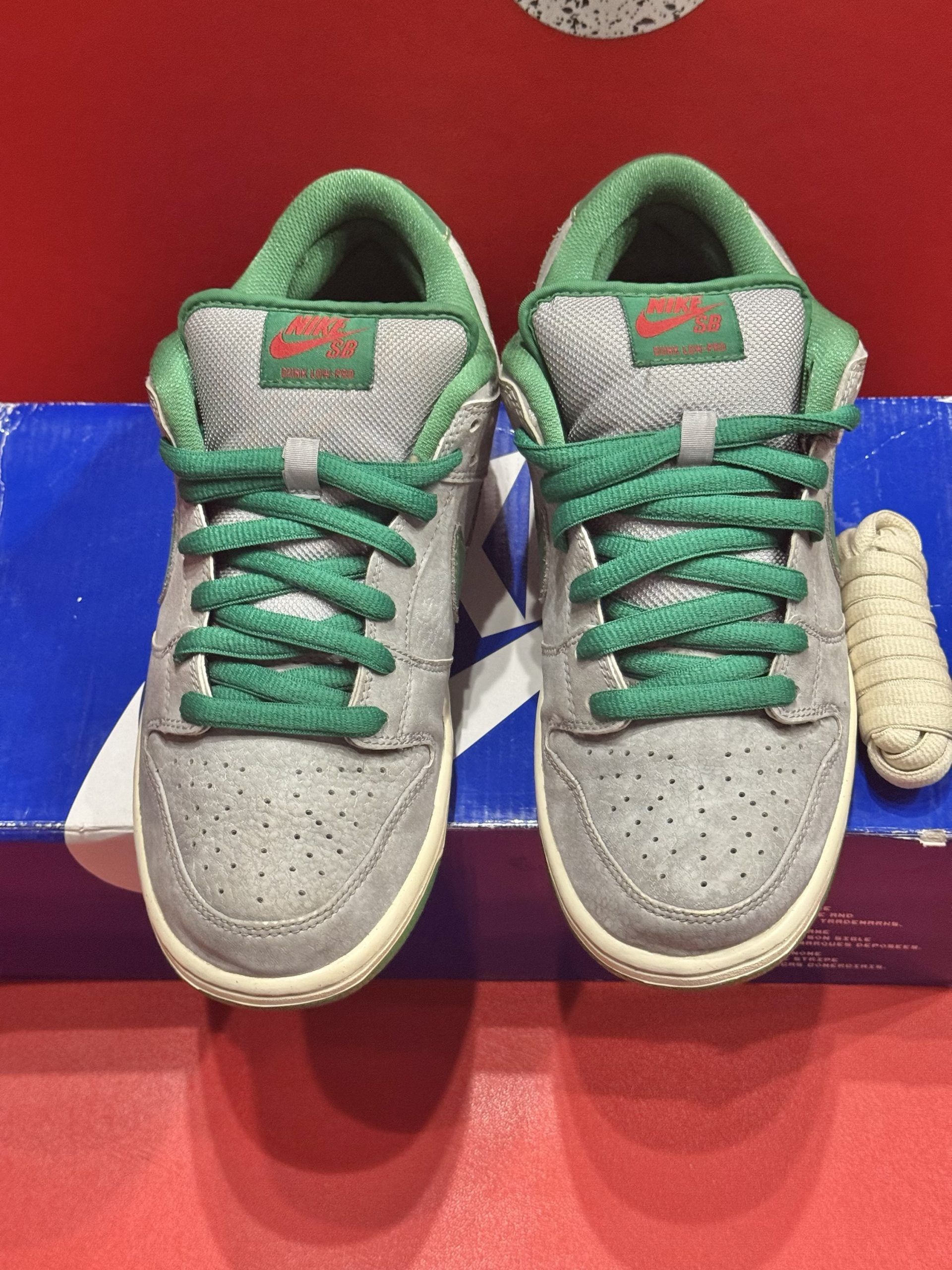 Nike SB Dunk Low sneakers in gray, green, and cream, displayed with the box and a red background.