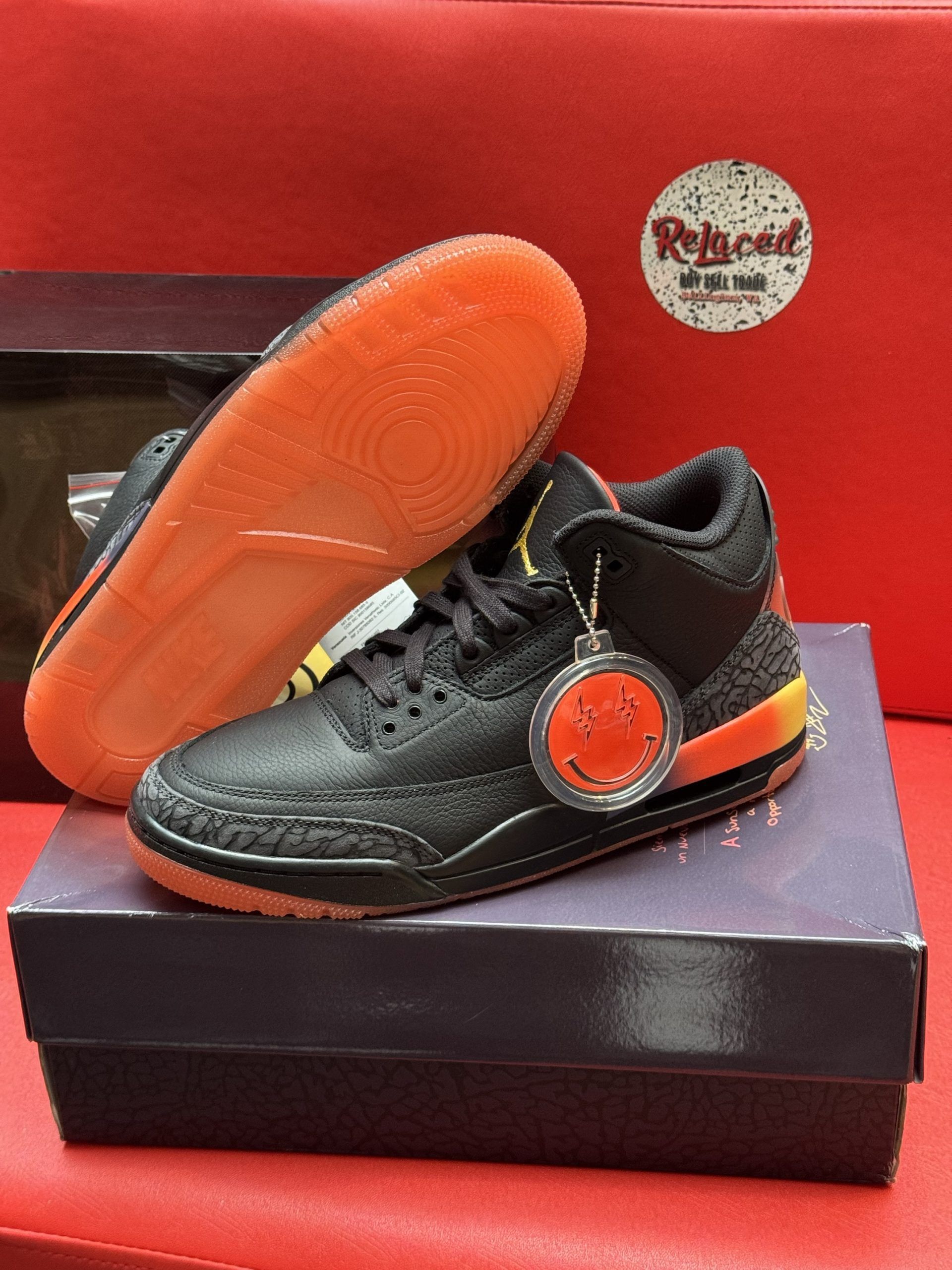 Black and orange Air Jordan sneakers with smiley face charm, on a purple box, red background.