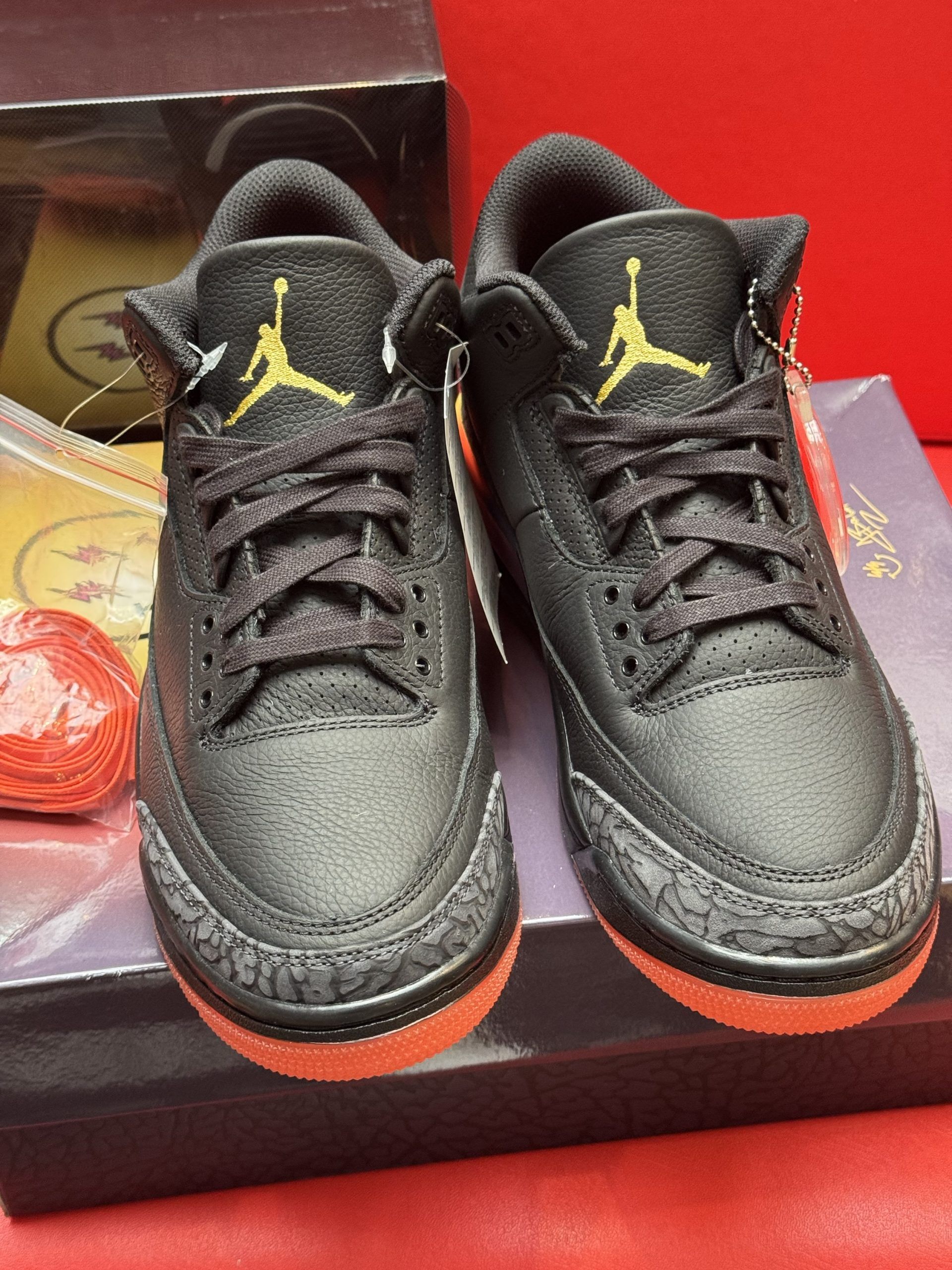 Black and orange Air Jordan sneakers with smiley face charm, on a purple box, red background.