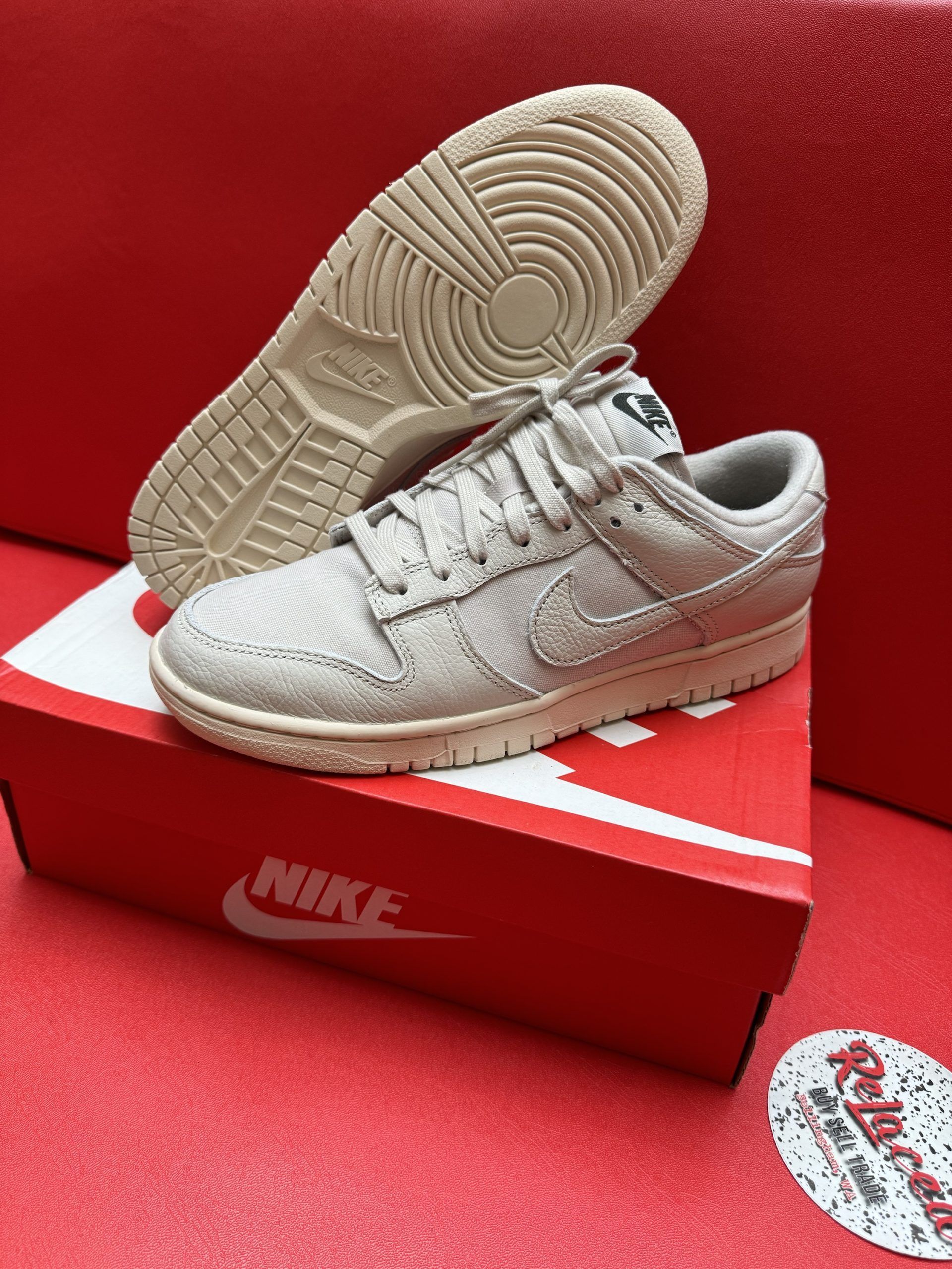 Beige Nike Dunk Low sneakers, one on top of the box, sole facing up. Red background.
