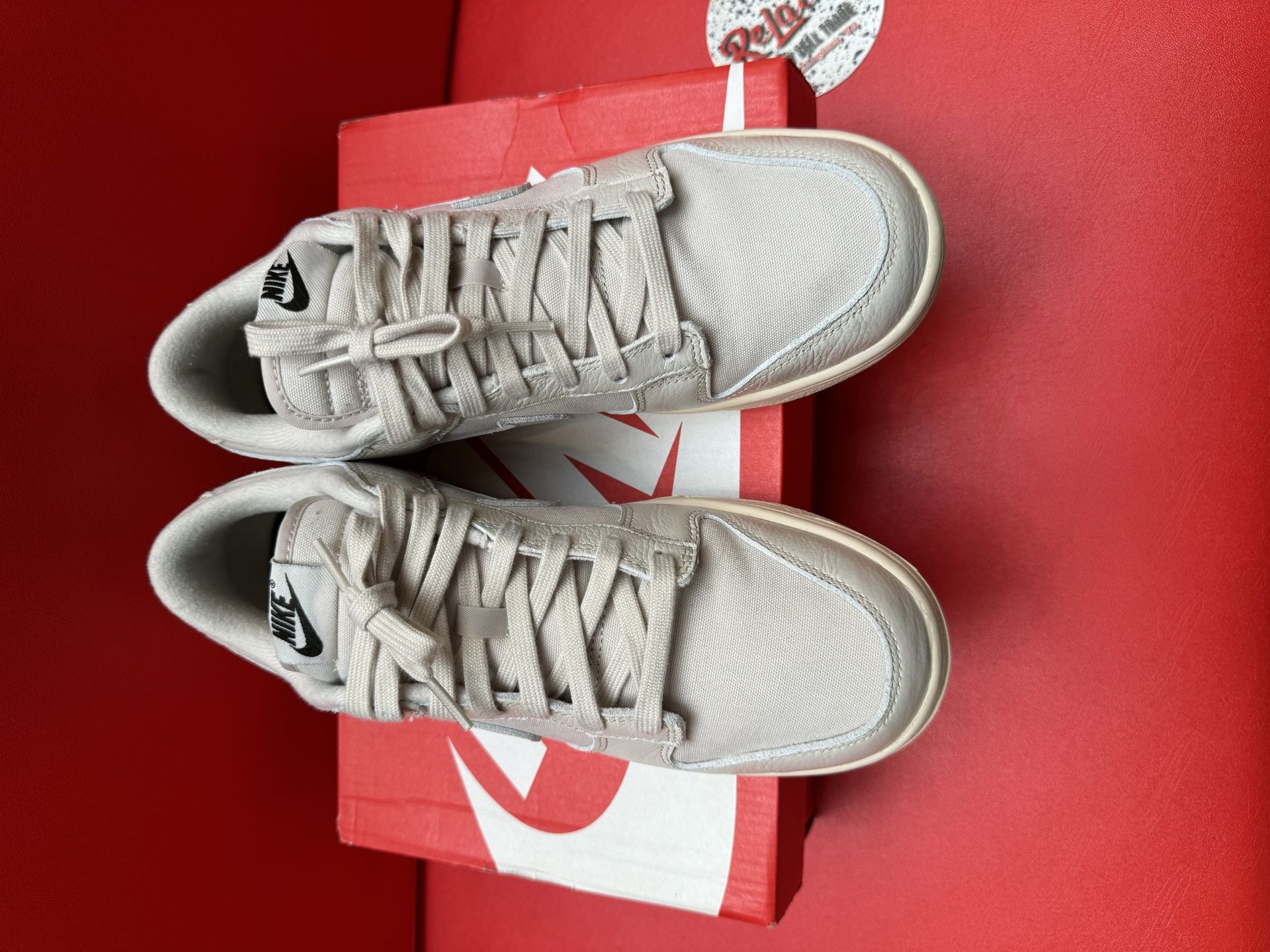 Beige Nike Dunk Low sneakers, one on top of the box, sole facing up. Red background.