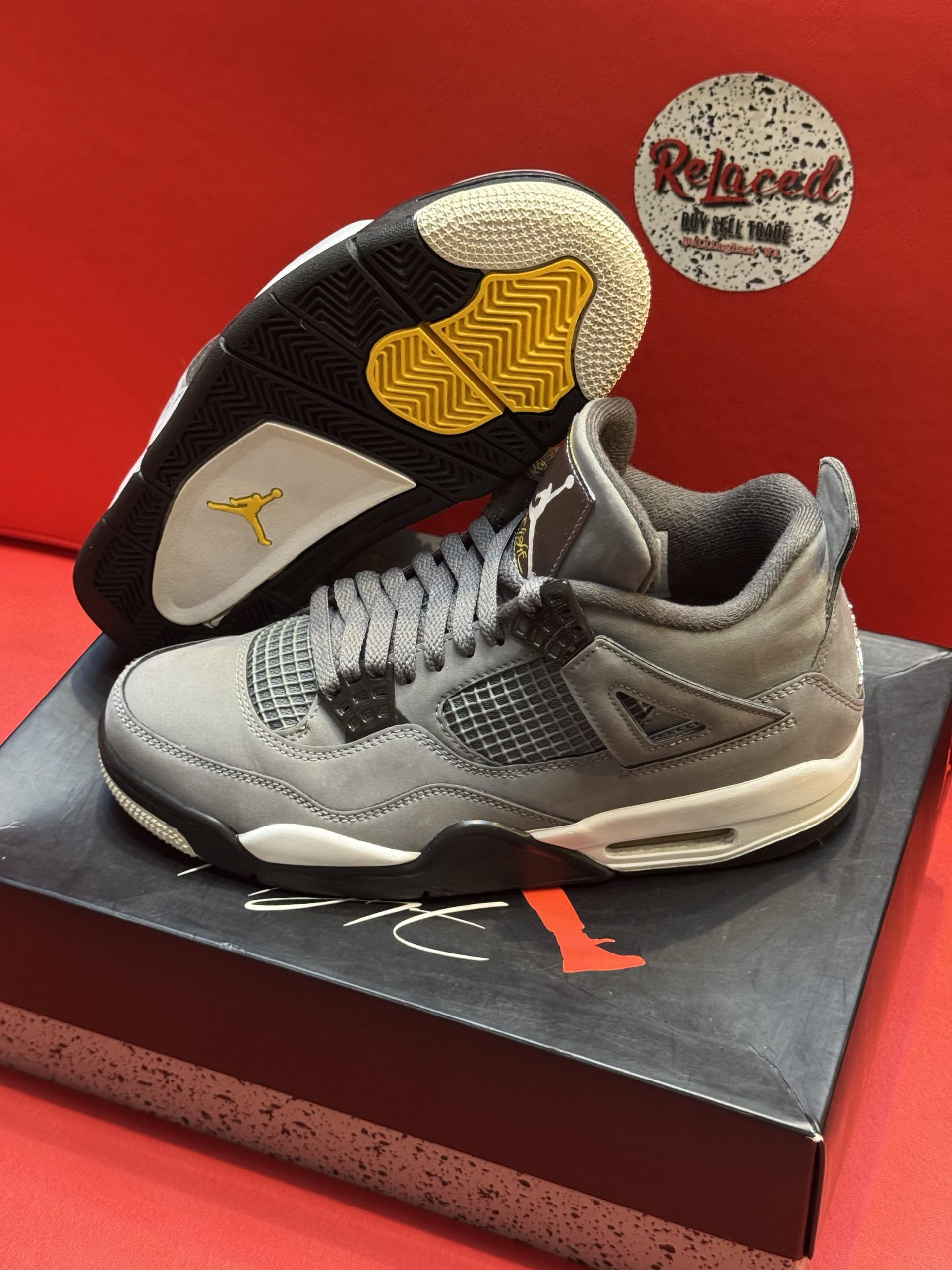 Gray Air Jordan 4 sneakers on a box, sole visible, against a red backdrop.