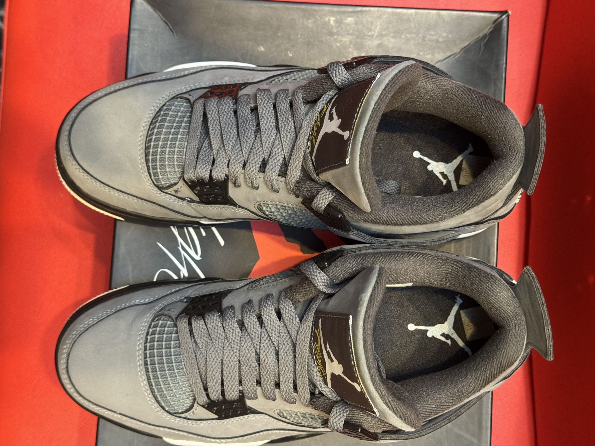 Gray Air Jordan 4 sneakers on a box, sole visible, against a red backdrop.