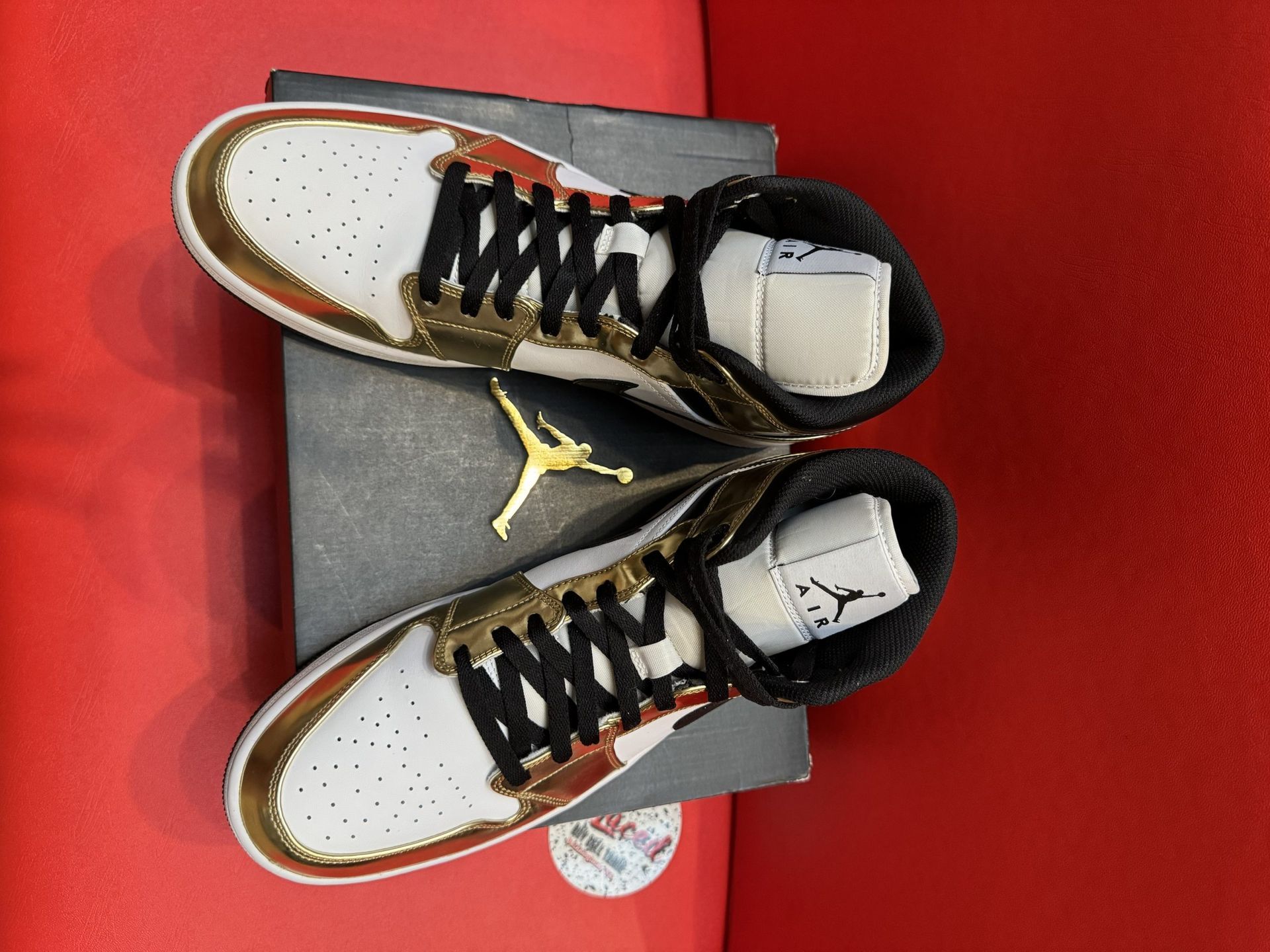 White, gold, and black Air Jordan 1 Mid sneaker on black box; black sole visible.