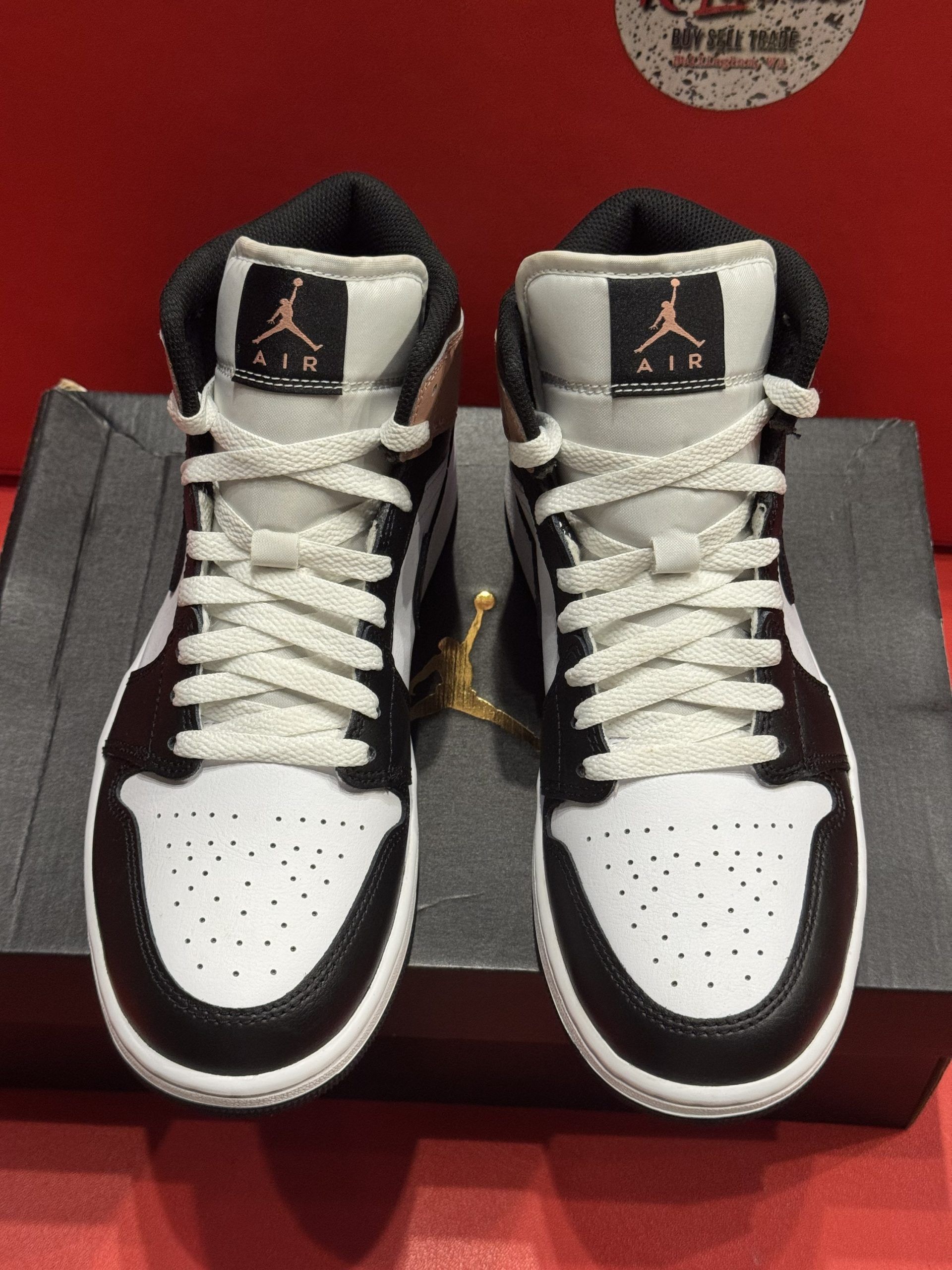 Air Jordan 1 Mid sneaker with a black, white, and tan color scheme, sitting on a black box, with the sole visible.