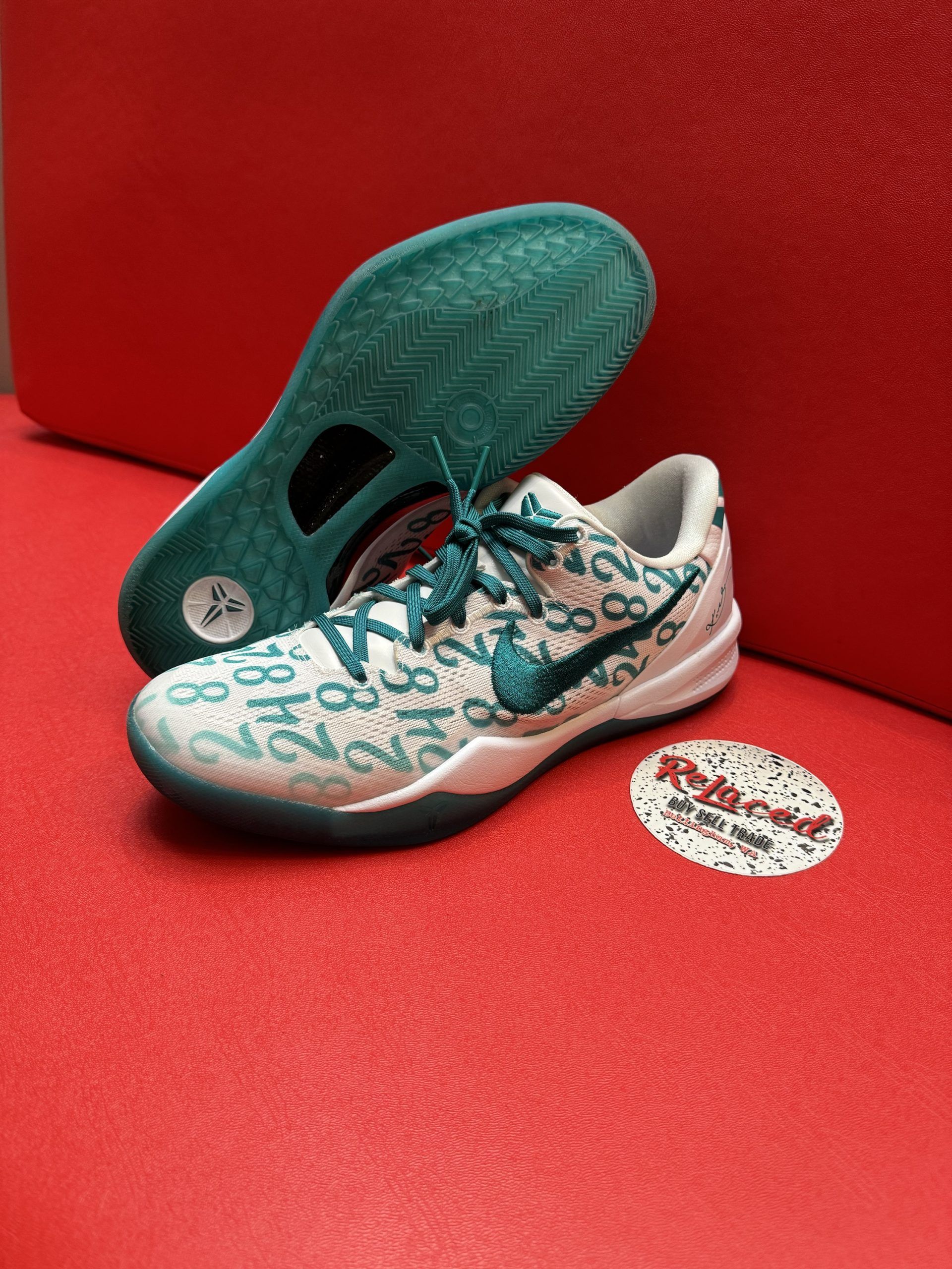 White and teal Nike basketball shoe with teal sole, number details, on a red surface.