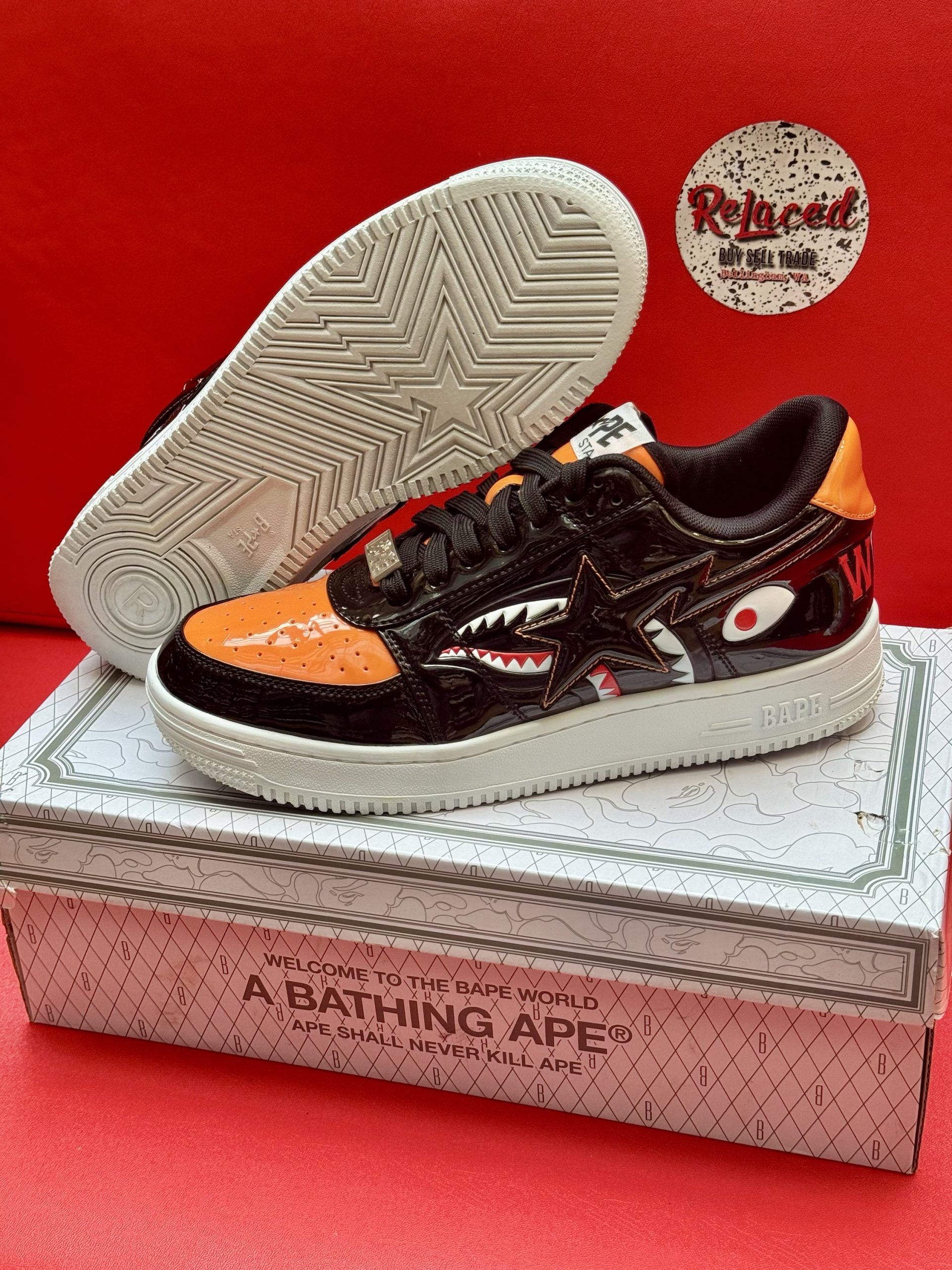 Black and orange BAPE sneakers with shark graphics, sitting on a branded box with a red backdrop.