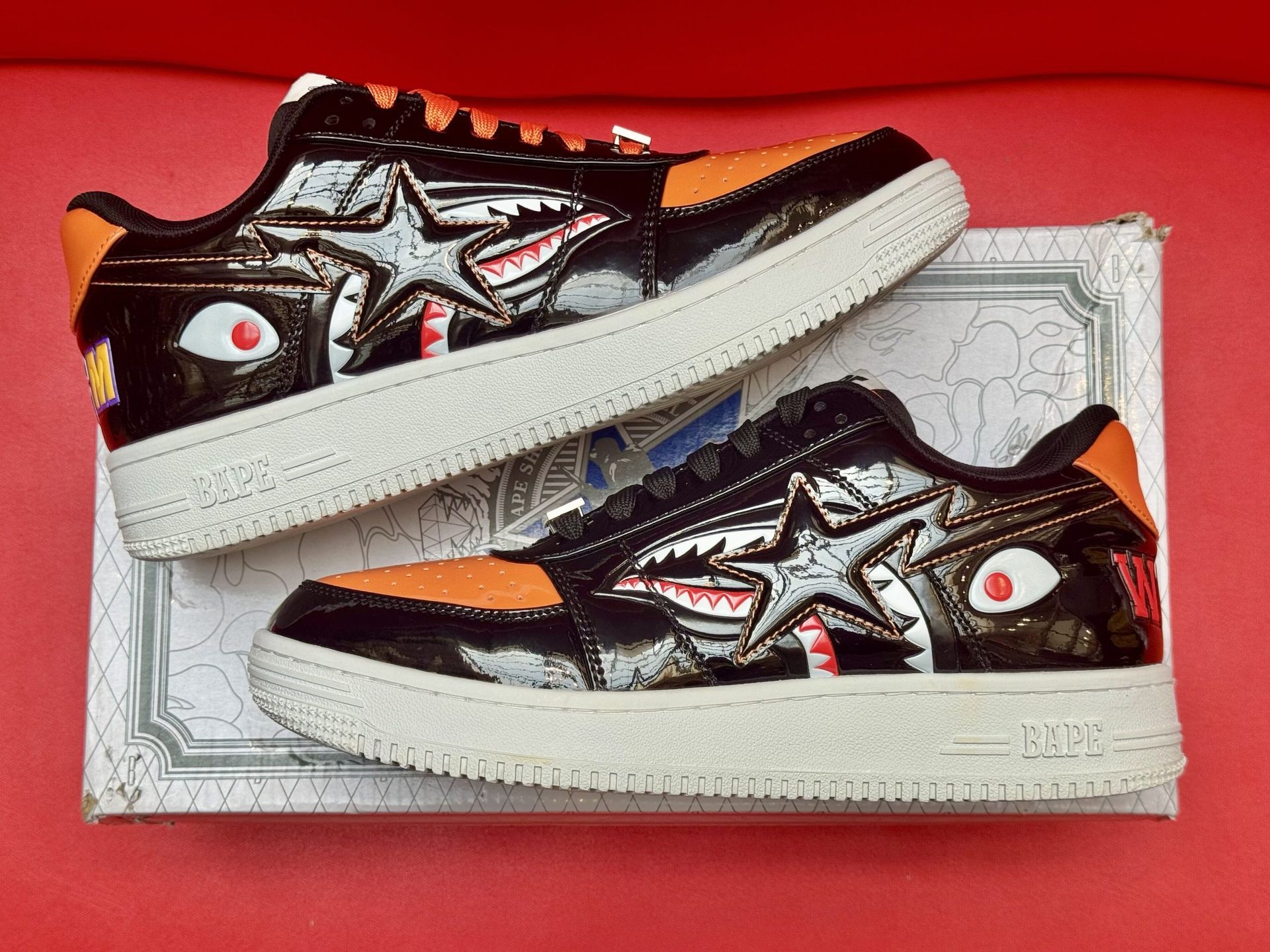 Black and orange BAPE sneakers with shark graphics, sitting on a branded box with a red backdrop.