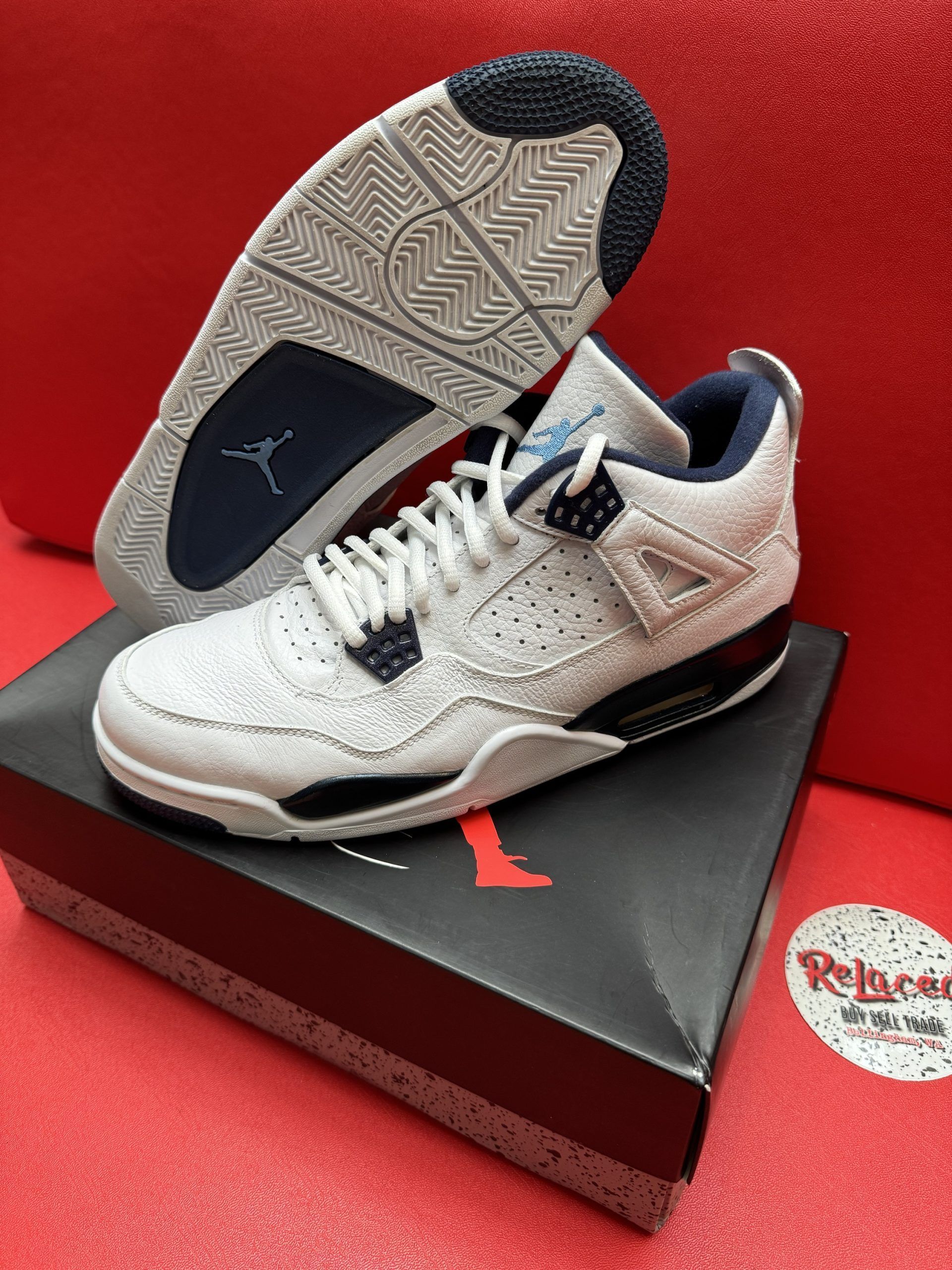 White and blue Air Jordan 4 sneakers on a shoebox, one turned to show the sole, on a red surface.