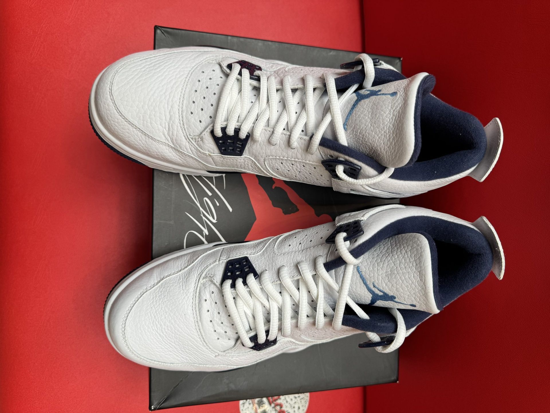 White and blue Air Jordan 4 sneakers on a shoebox, one turned to show the sole, on a red surface.