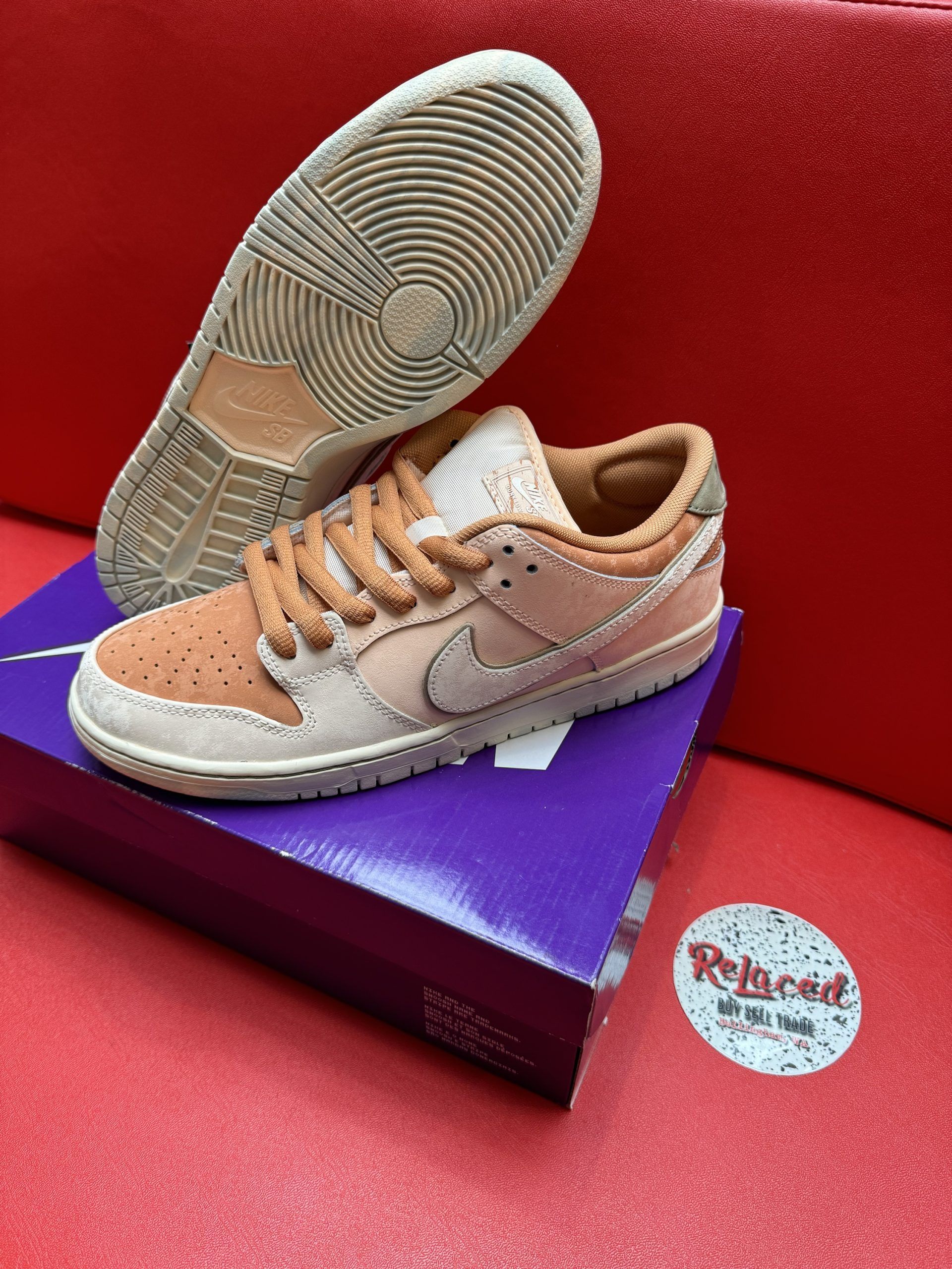 Tan and cream Nike Dunk SB sneakers on a purple box, with one turned over to show sole. Red background.
