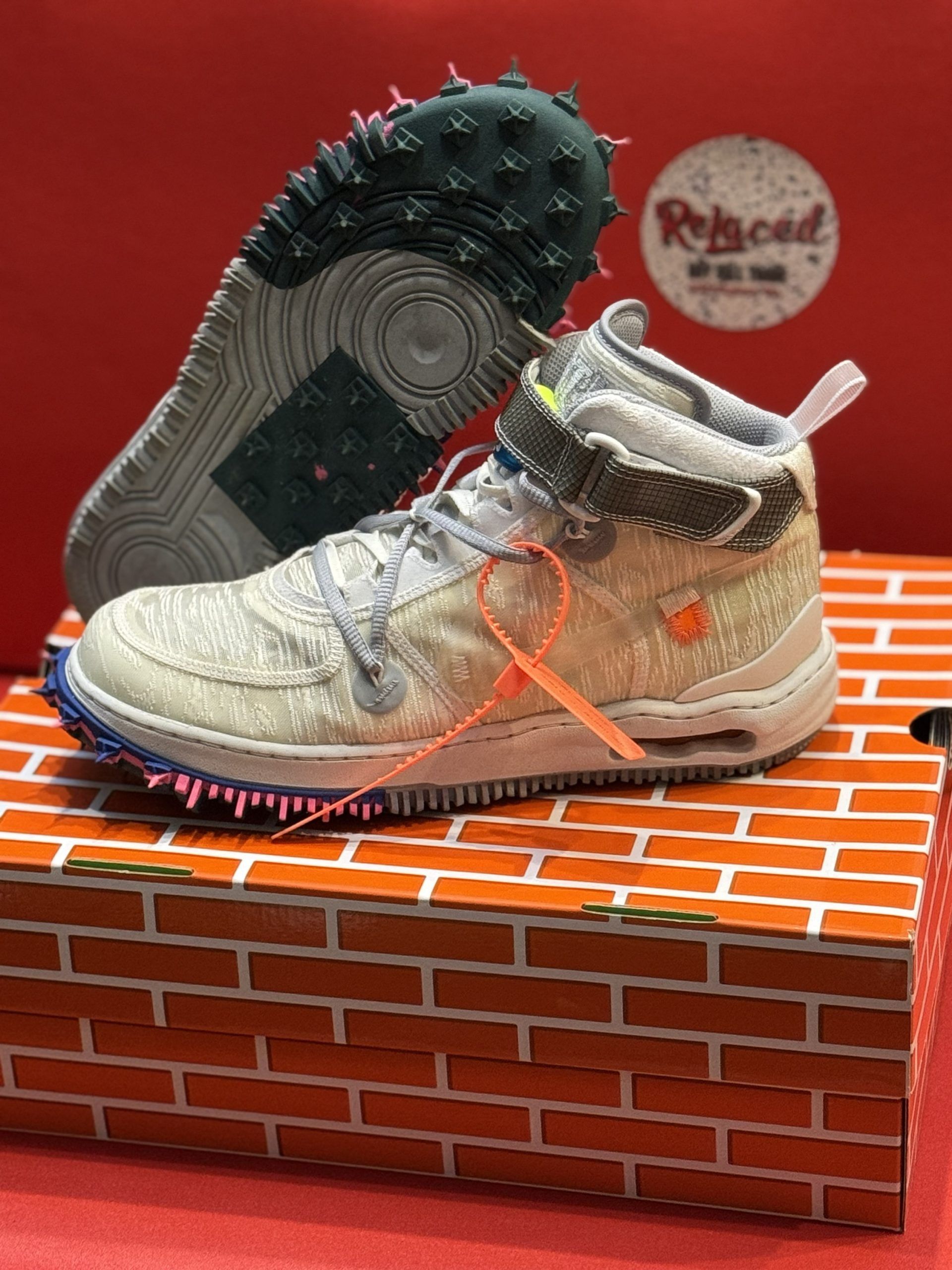 Tan Nike high-top sneakers with spiky soles and orange tag, on a brick-patterned box.