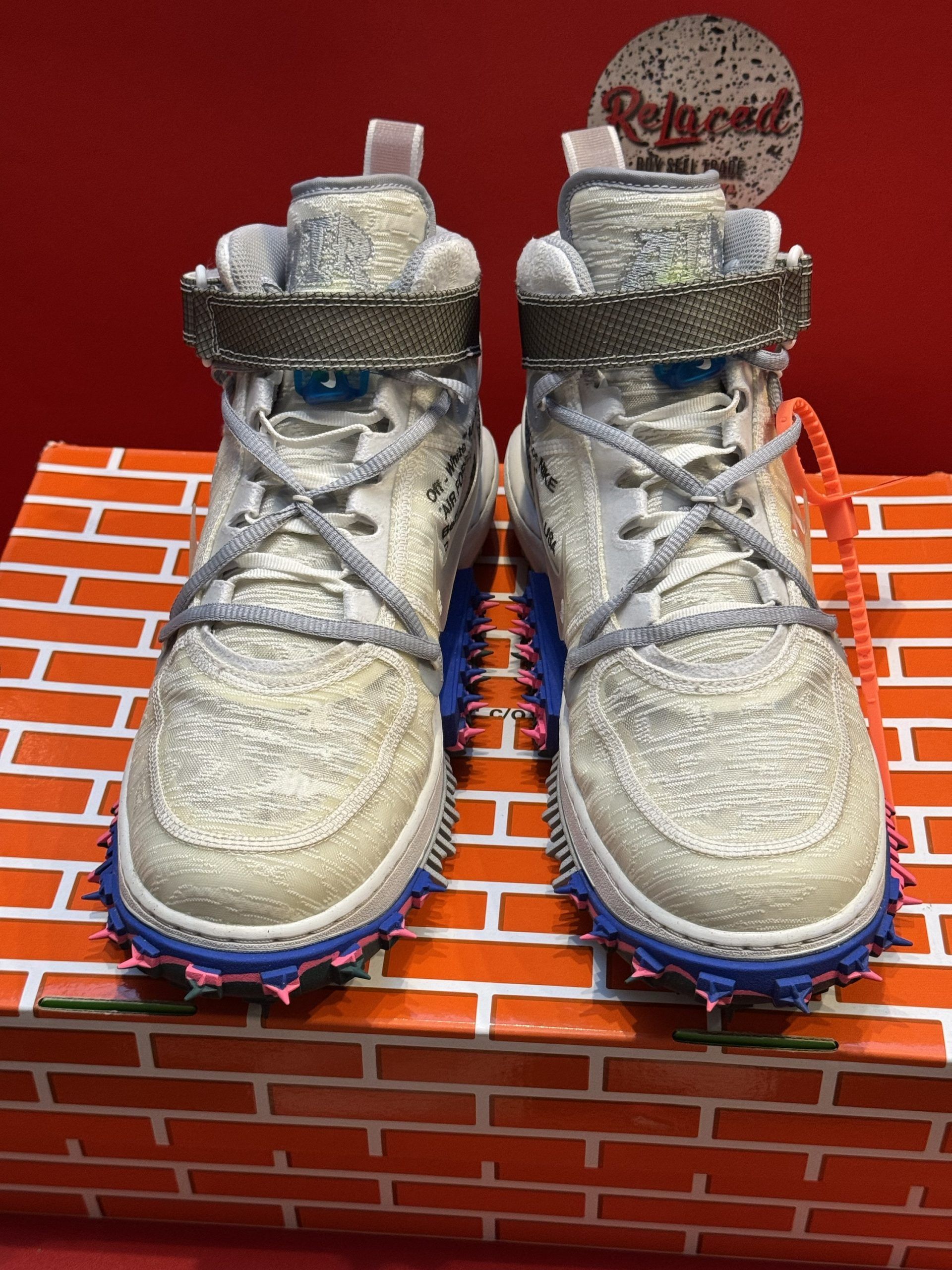 Tan Nike high-top sneakers with spiky soles and orange tag, on a brick-patterned box.