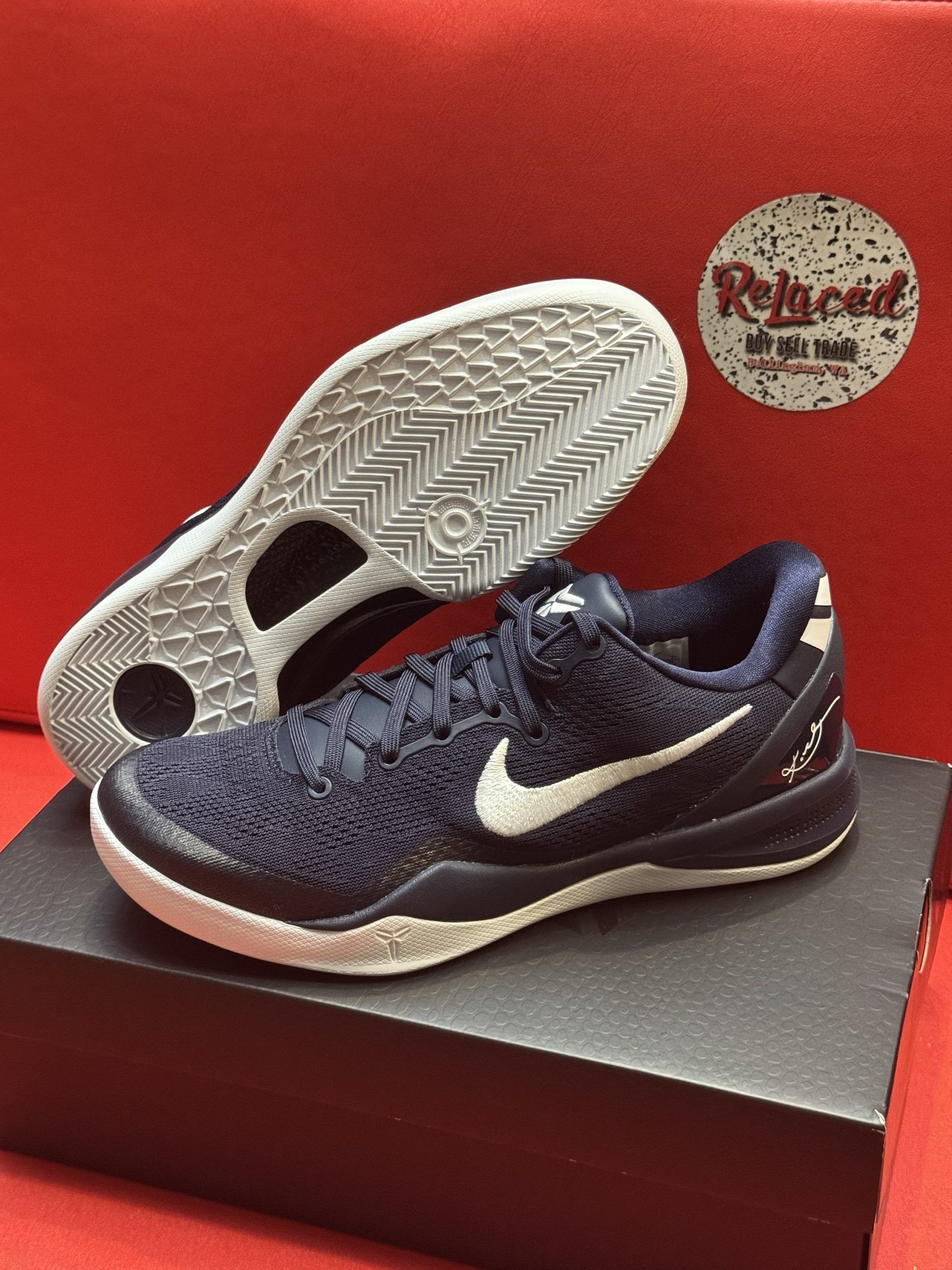 Navy blue Nike basketball shoes with white accents, one on top of the other, on a black box, with a red background.