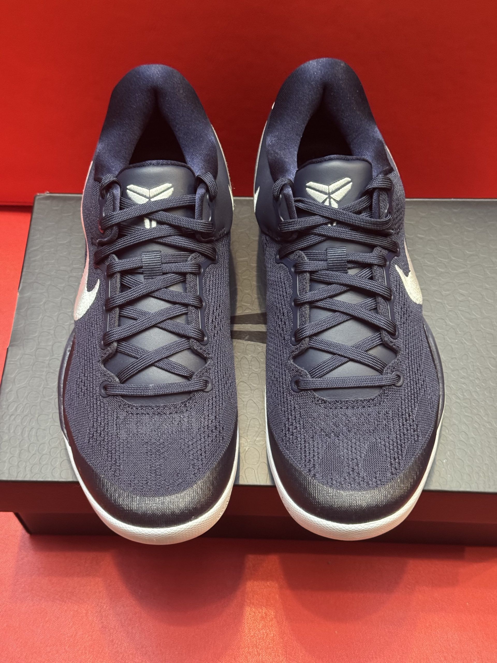 Navy blue Nike basketball shoes with white accents, one on top of the other, on a black box, with a red background.
