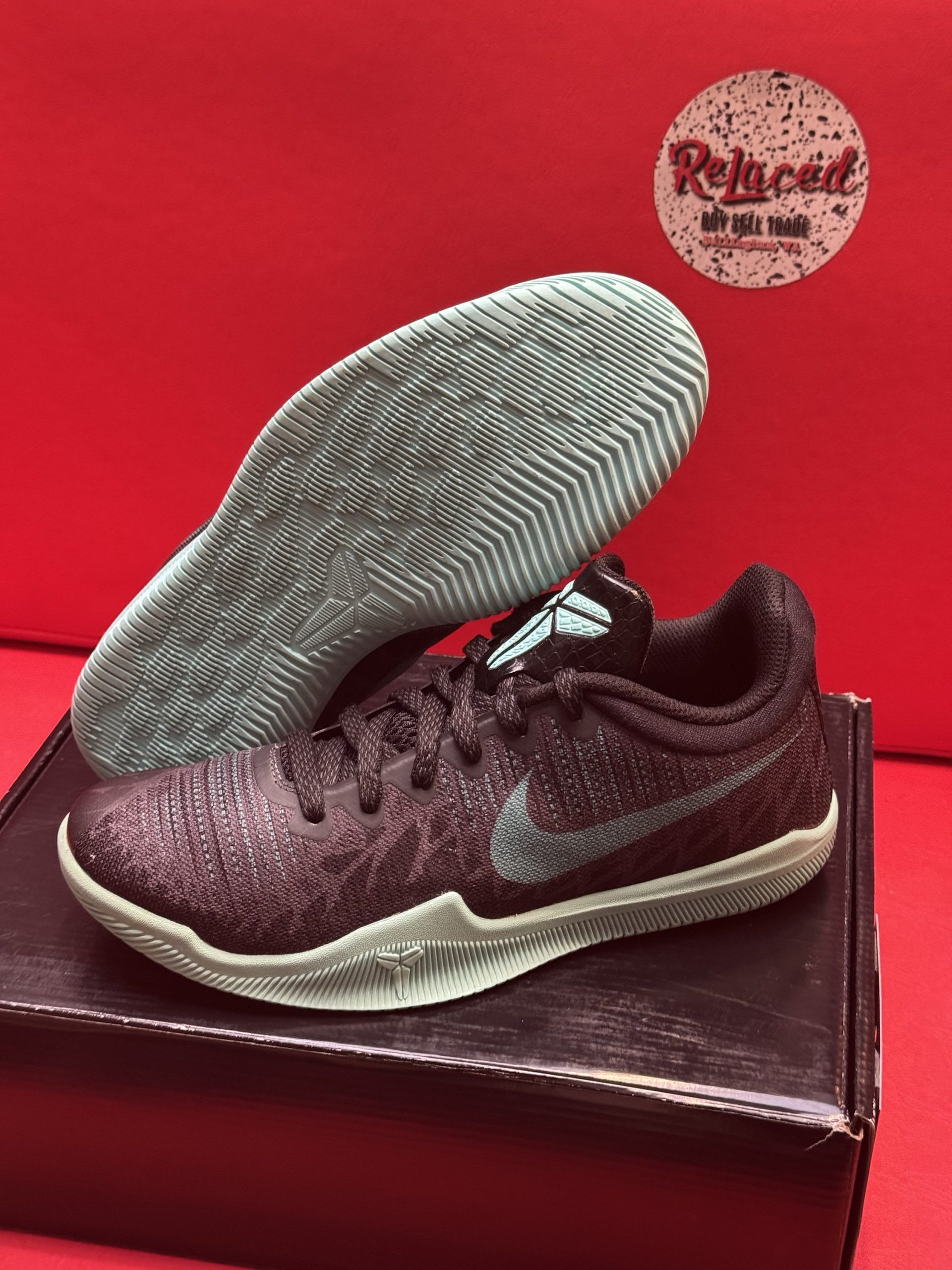 Black and gray Kobe basketball sneaker with matching sole, set on a box against a red backdrop.