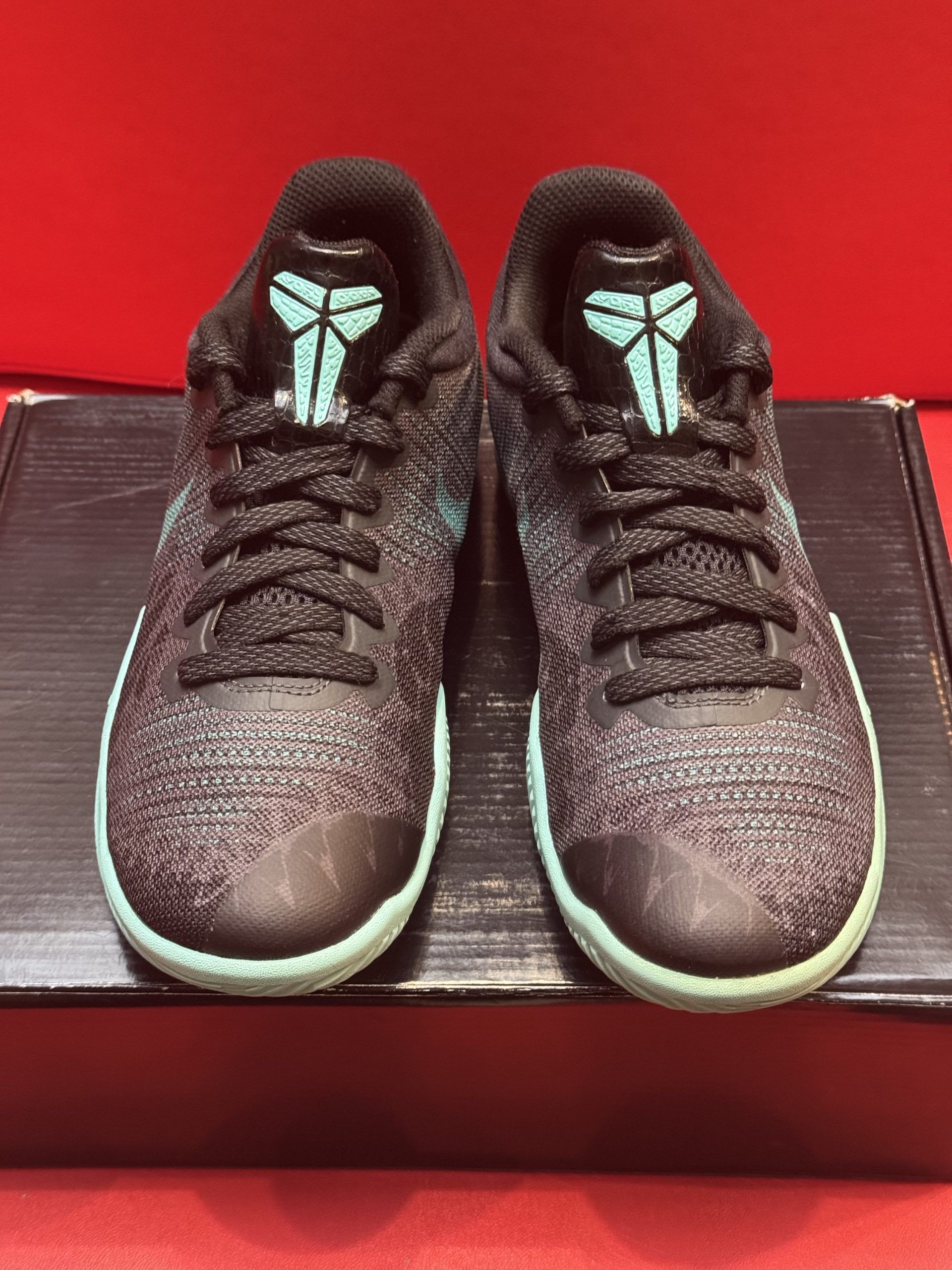 Black and gray Kobe basketball sneaker with matching sole, set on a box against a red backdrop.