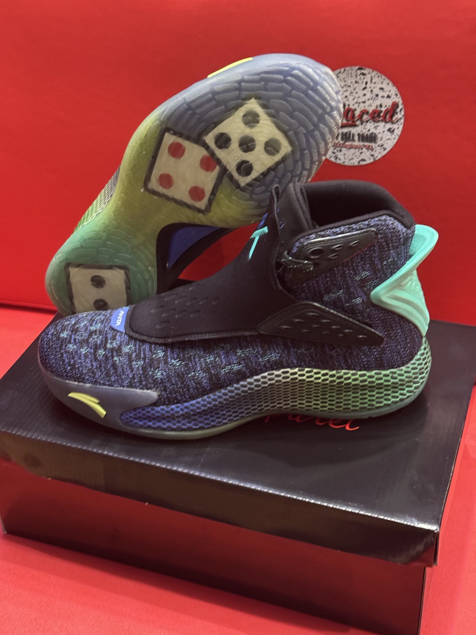 Blue and green basketball shoe with dice-pattern sole. Black strap and collar. Sits on a black box, red background.
