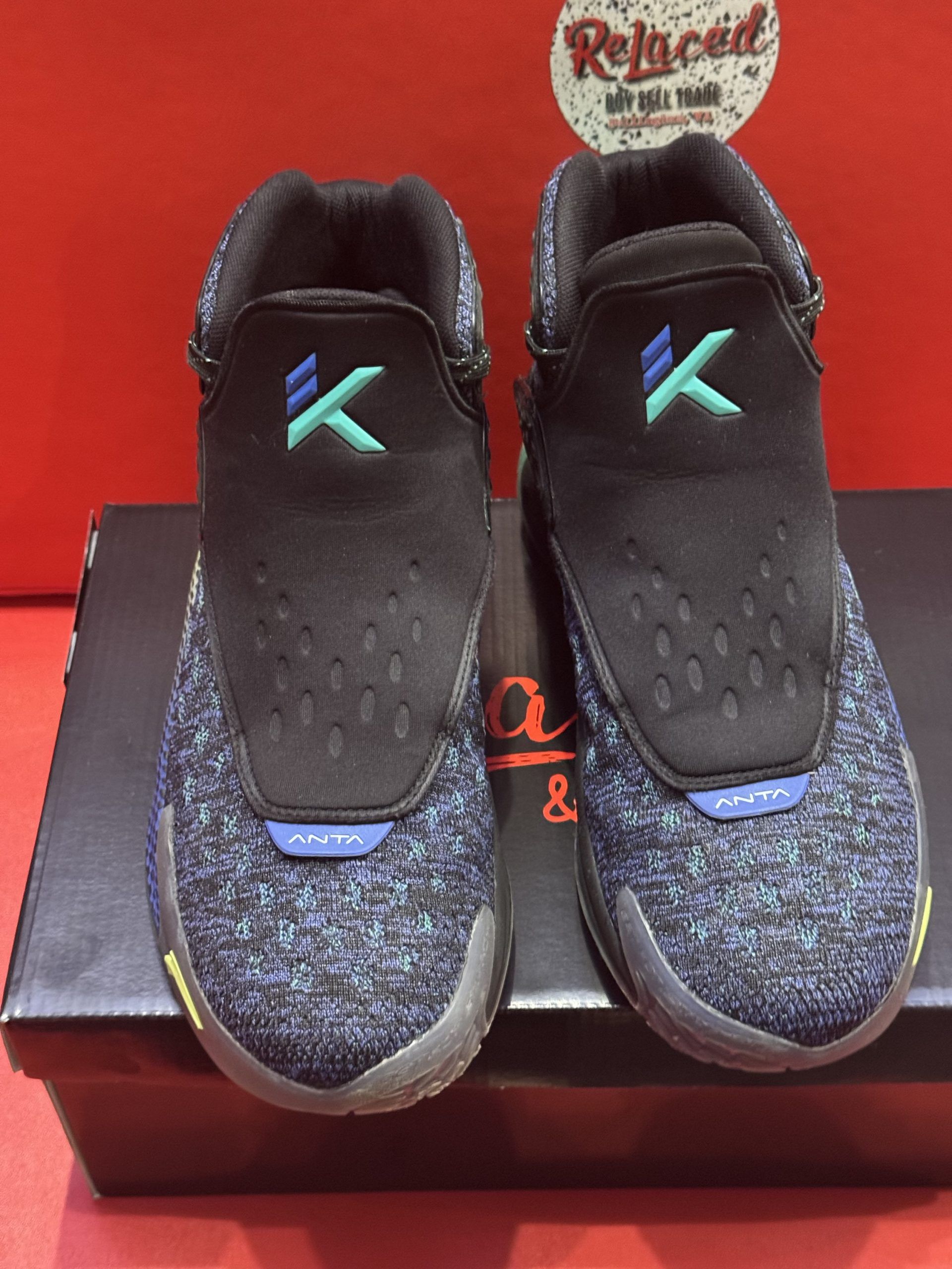 Blue and green basketball shoe with dice-pattern sole. Black strap and collar. Sits on a black box, red background.