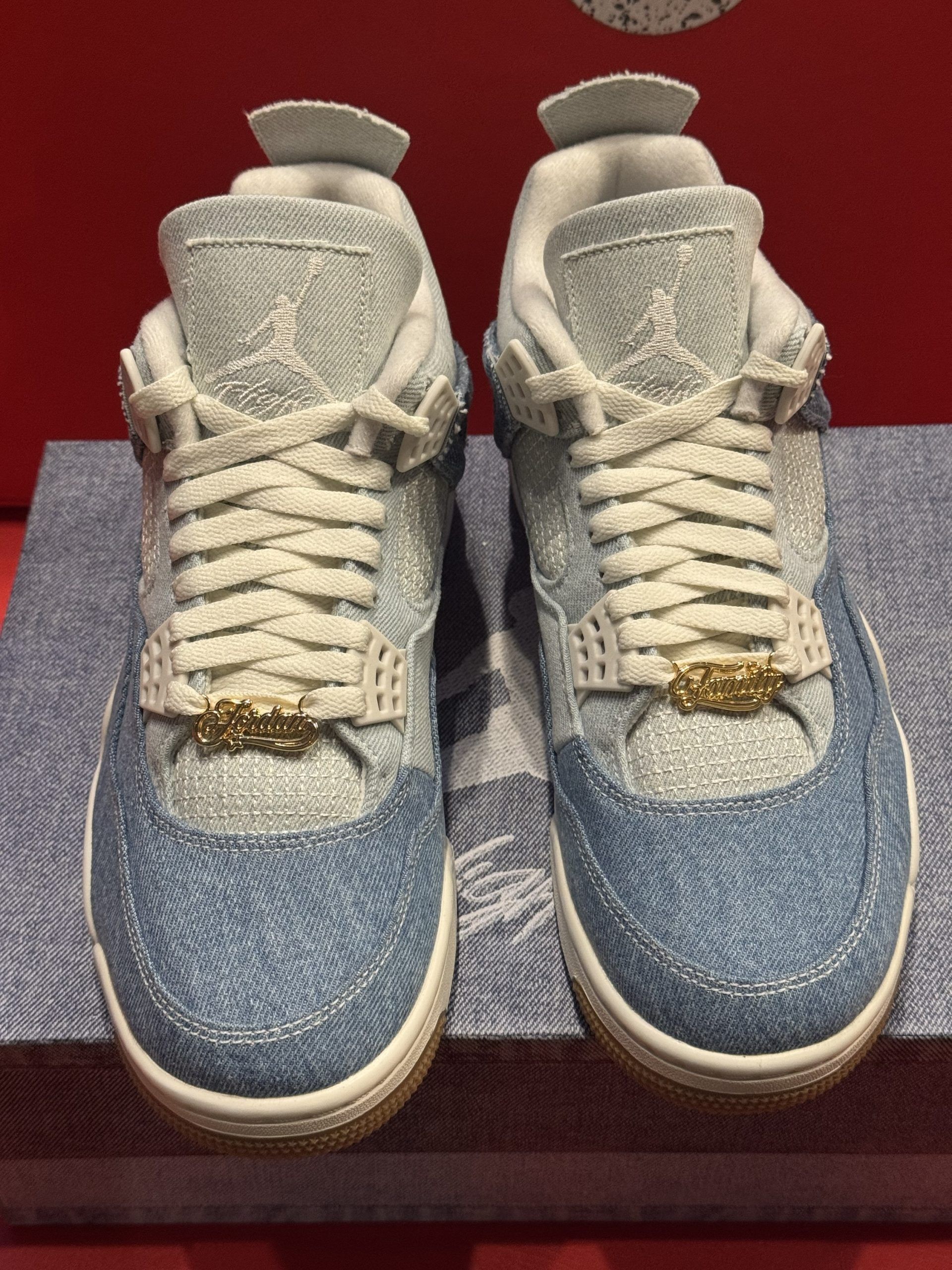 Air Jordan 4 sneakers with denim upper and brown soles, on denim box, against a red background.