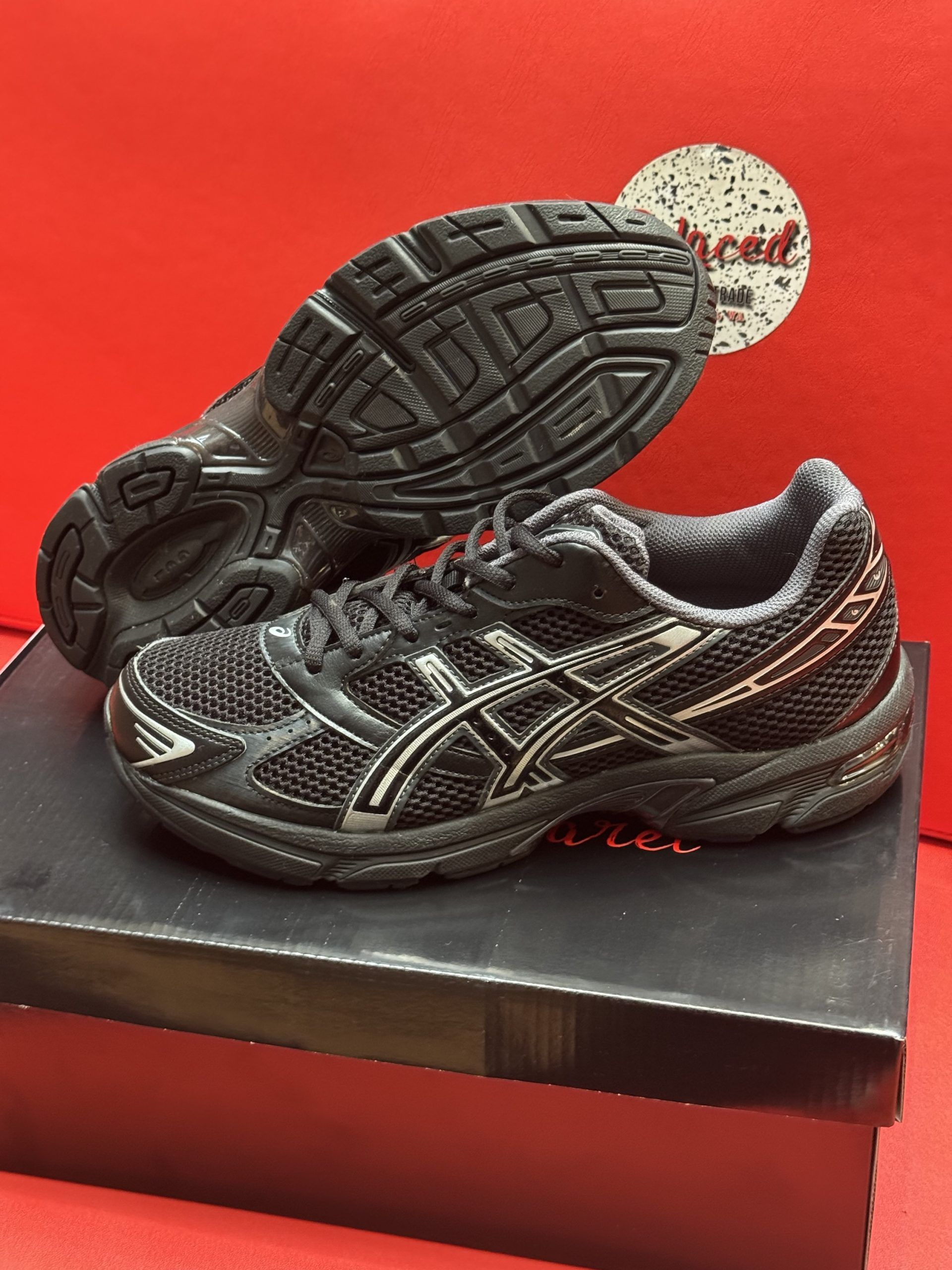Black ASICS running shoe on a black box, with the sole visible. Red background.