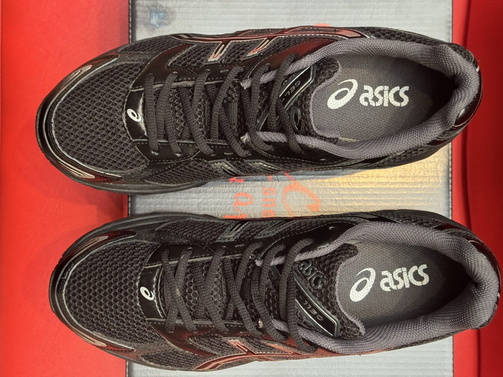 Black ASICS running shoe on a black box, with the sole visible. Red background.