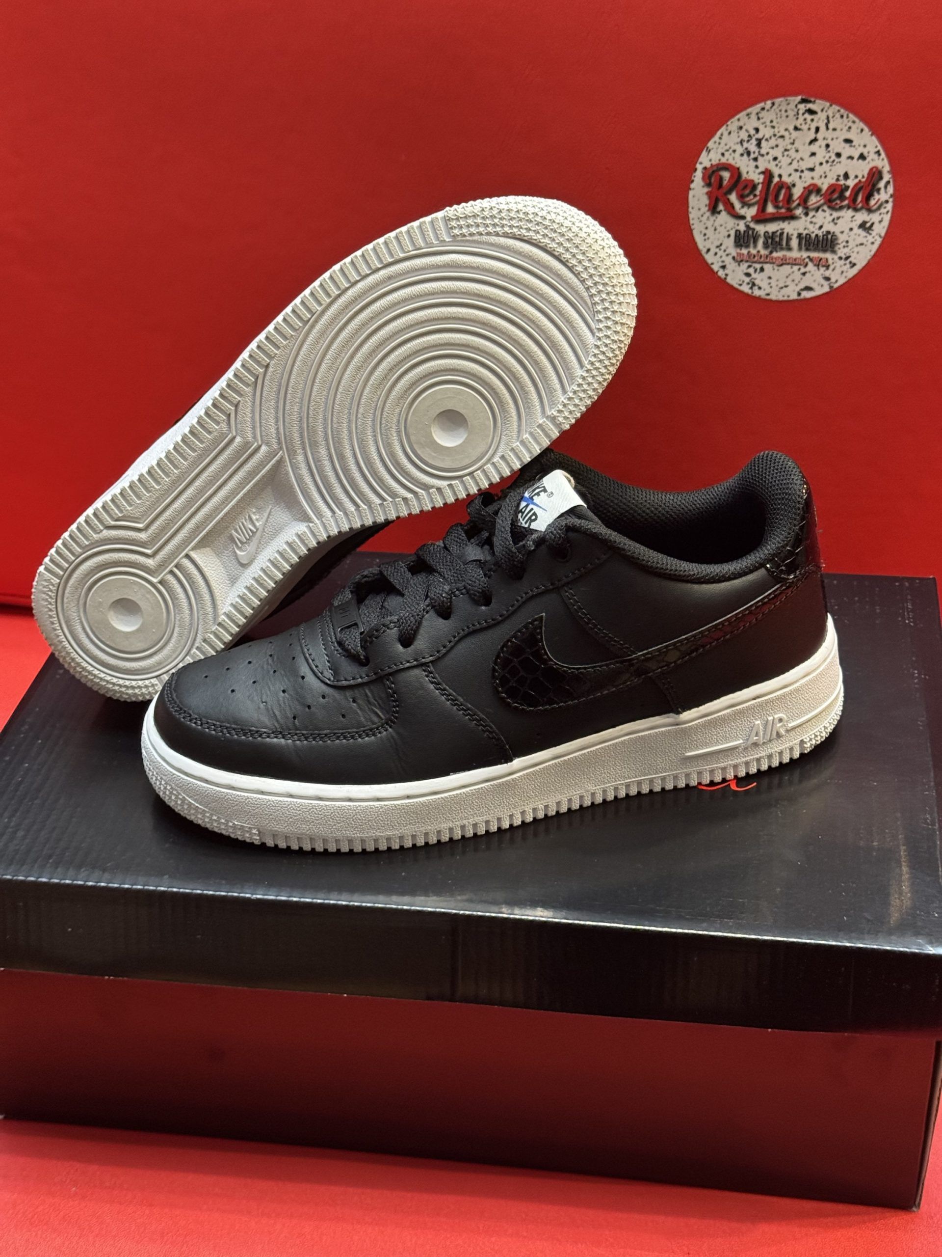 Black Nike Air Force 1 sneaker with a white speckled sole, set against a red background, displayed on a black box.