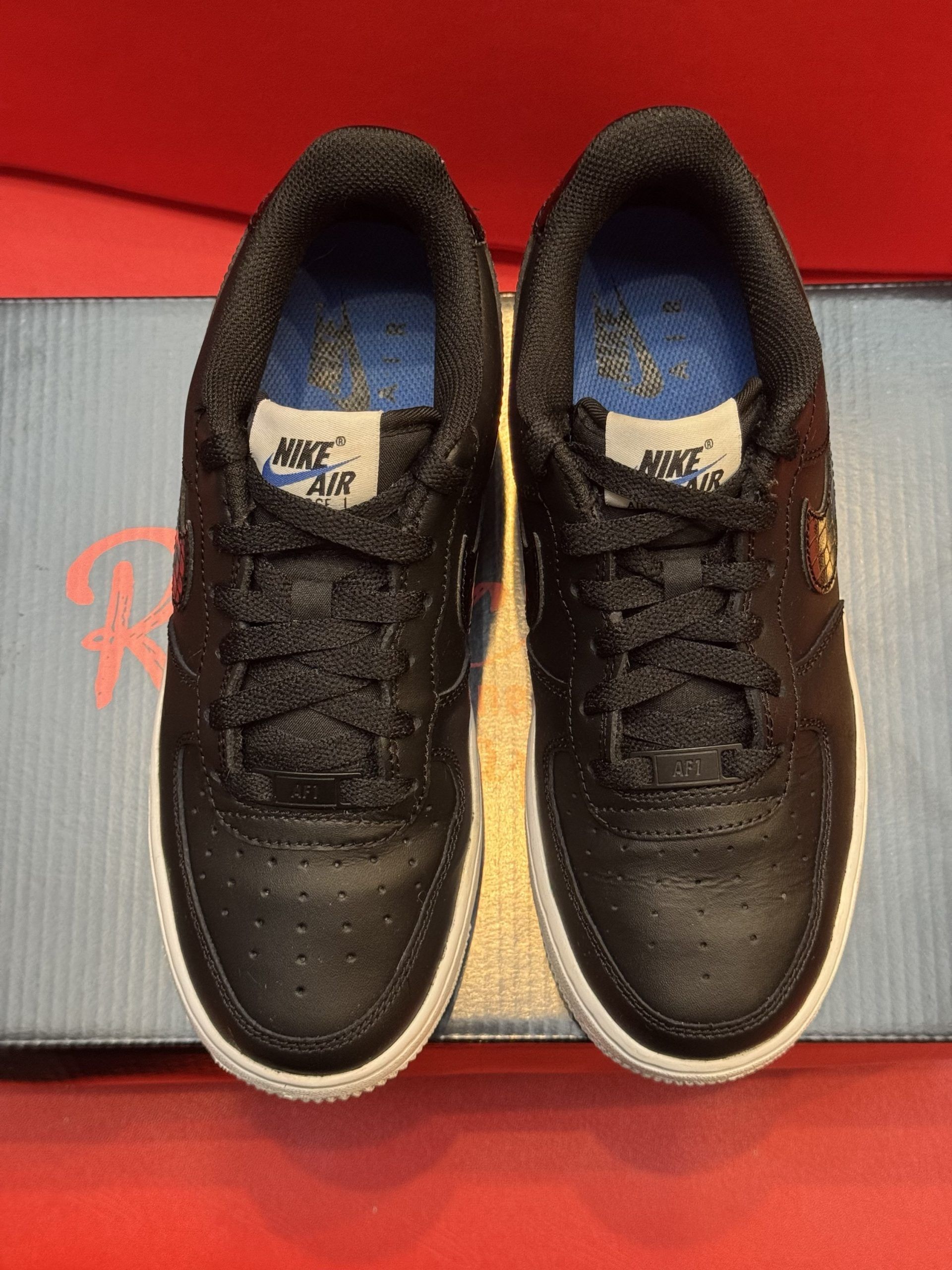 Black Nike Air Force 1 sneaker with a white speckled sole, set against a red background, displayed on a black box.