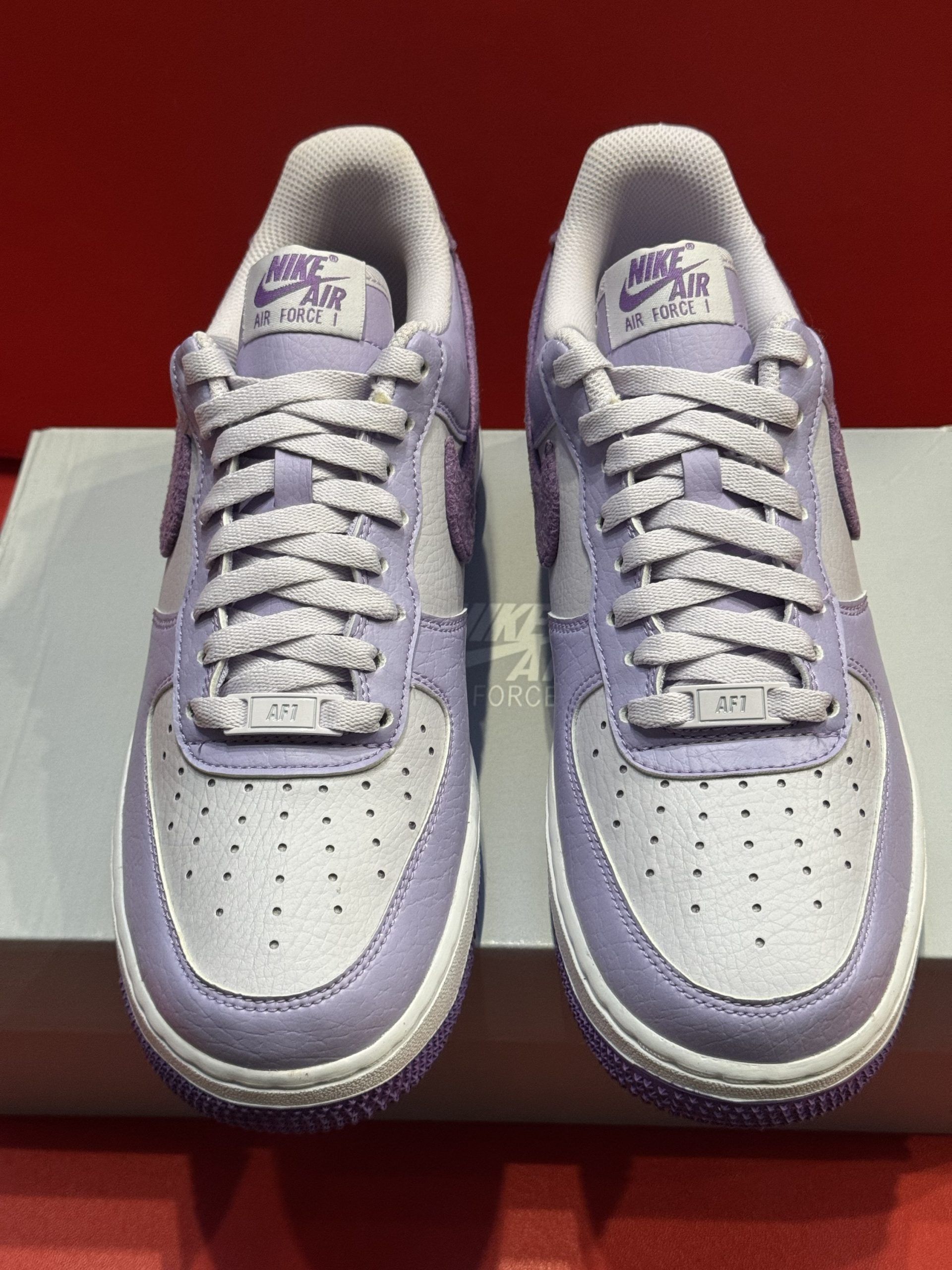 Purple and white Nike Air Force 1 sneakers on a white box, red background.