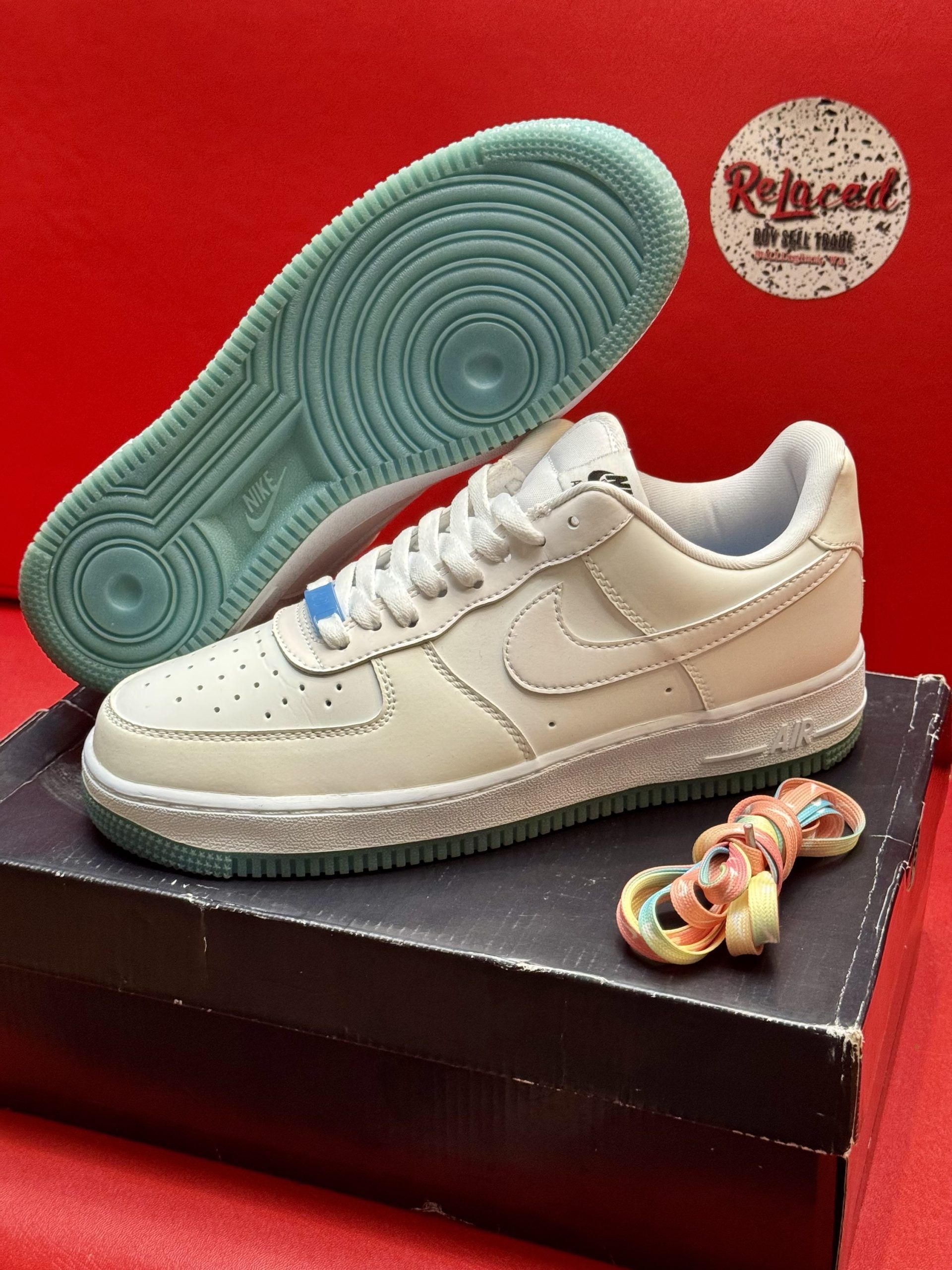 Pair of cream Nike Air Force 1 sneakers with blue soles on a shoebox, with a colorful pin.
