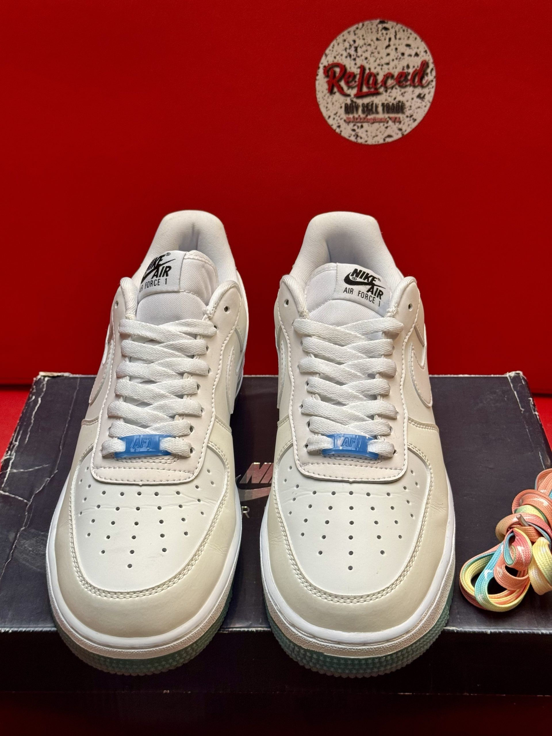Pair of cream Nike Air Force 1 sneakers with blue soles on a shoebox, with a colorful pin.