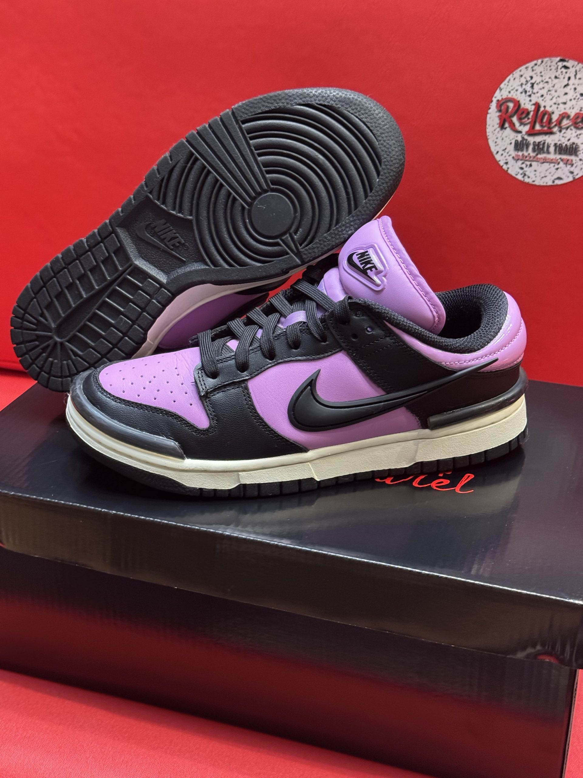 Black and purple Nike Dunk Low sneakers with black sole, resting on a shoebox against a red backdrop.