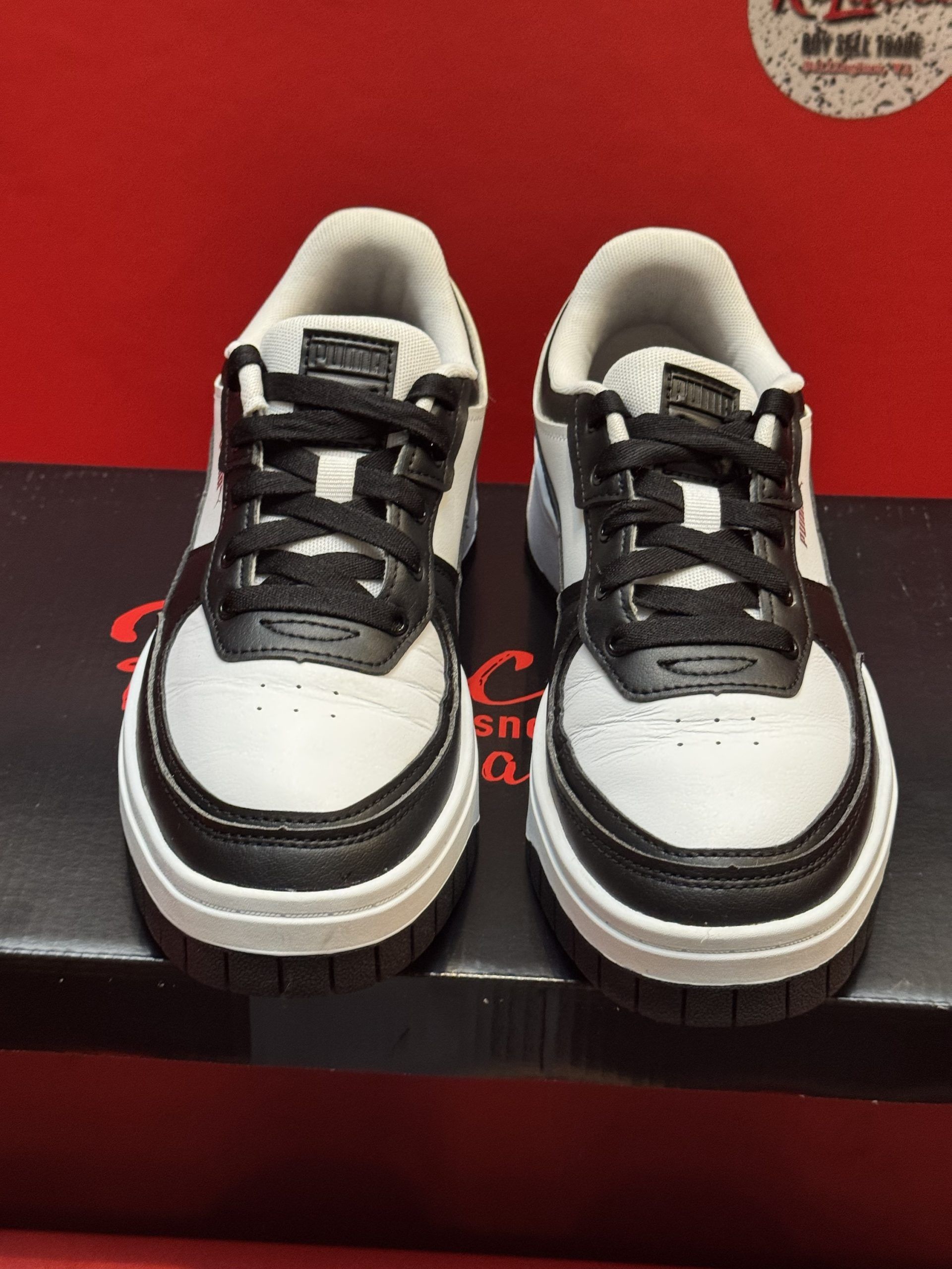 White and black sneakers with black sole, sitting on a black box against a red background.