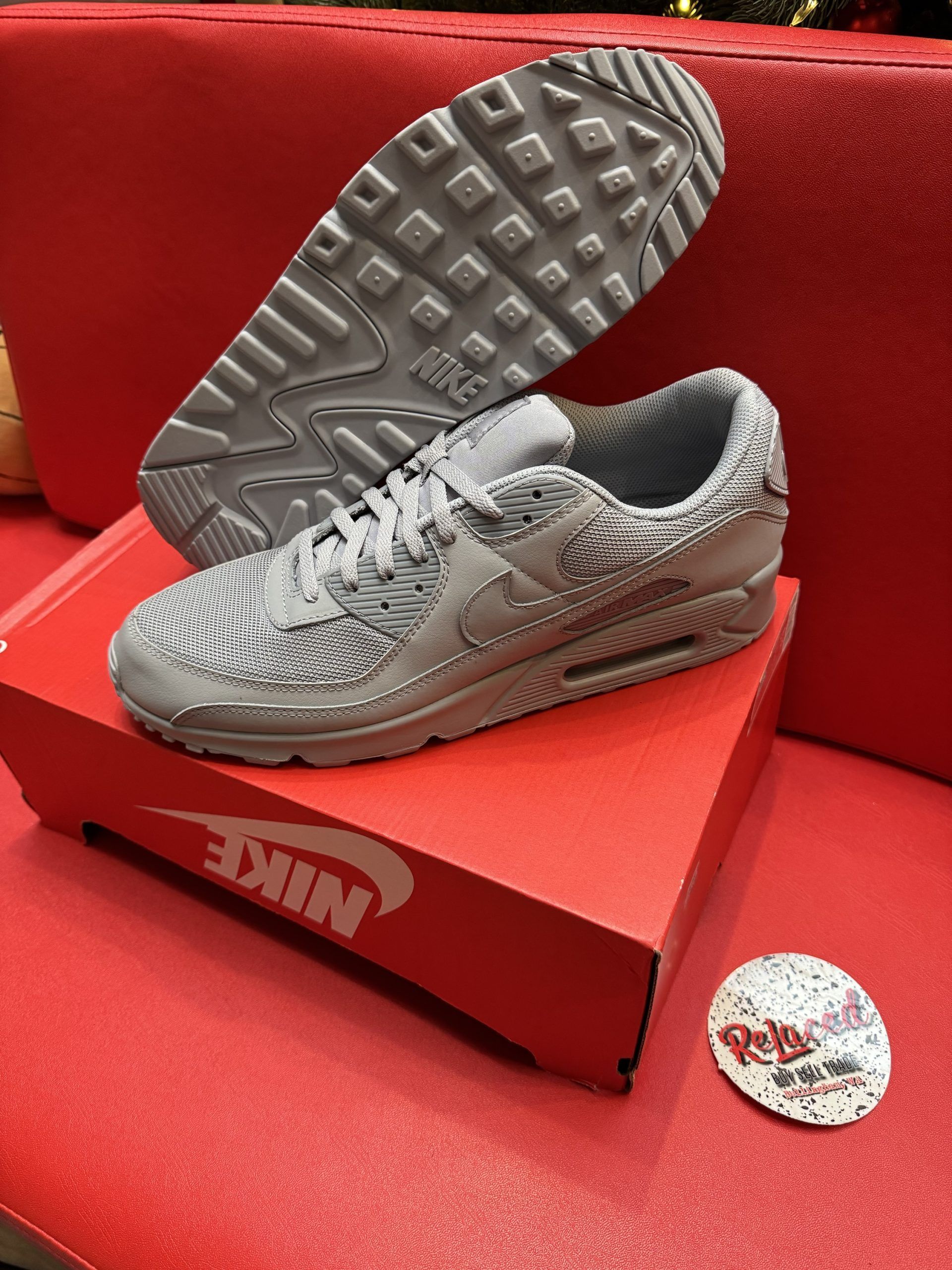 Gray Nike Air Max 90 sneakers on a red Nike box, with the sole visible. Red background.