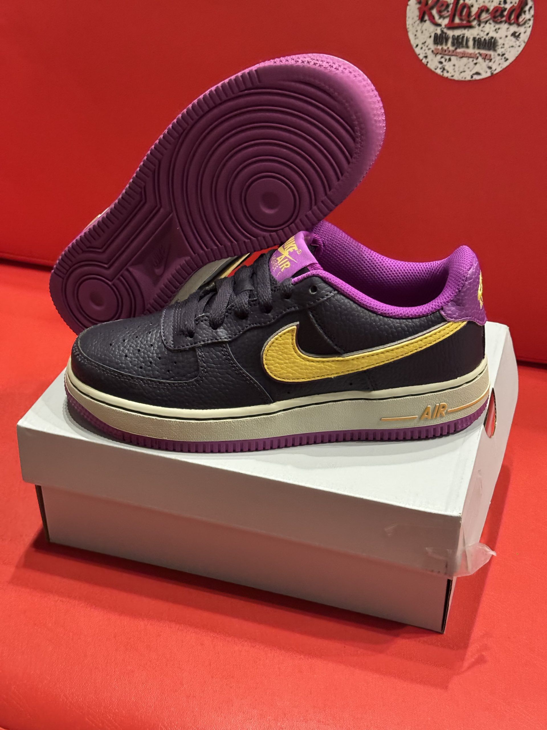 Black and yellow Nike Air Force 1 shoe on a white box, set against a red background. Purple sole.