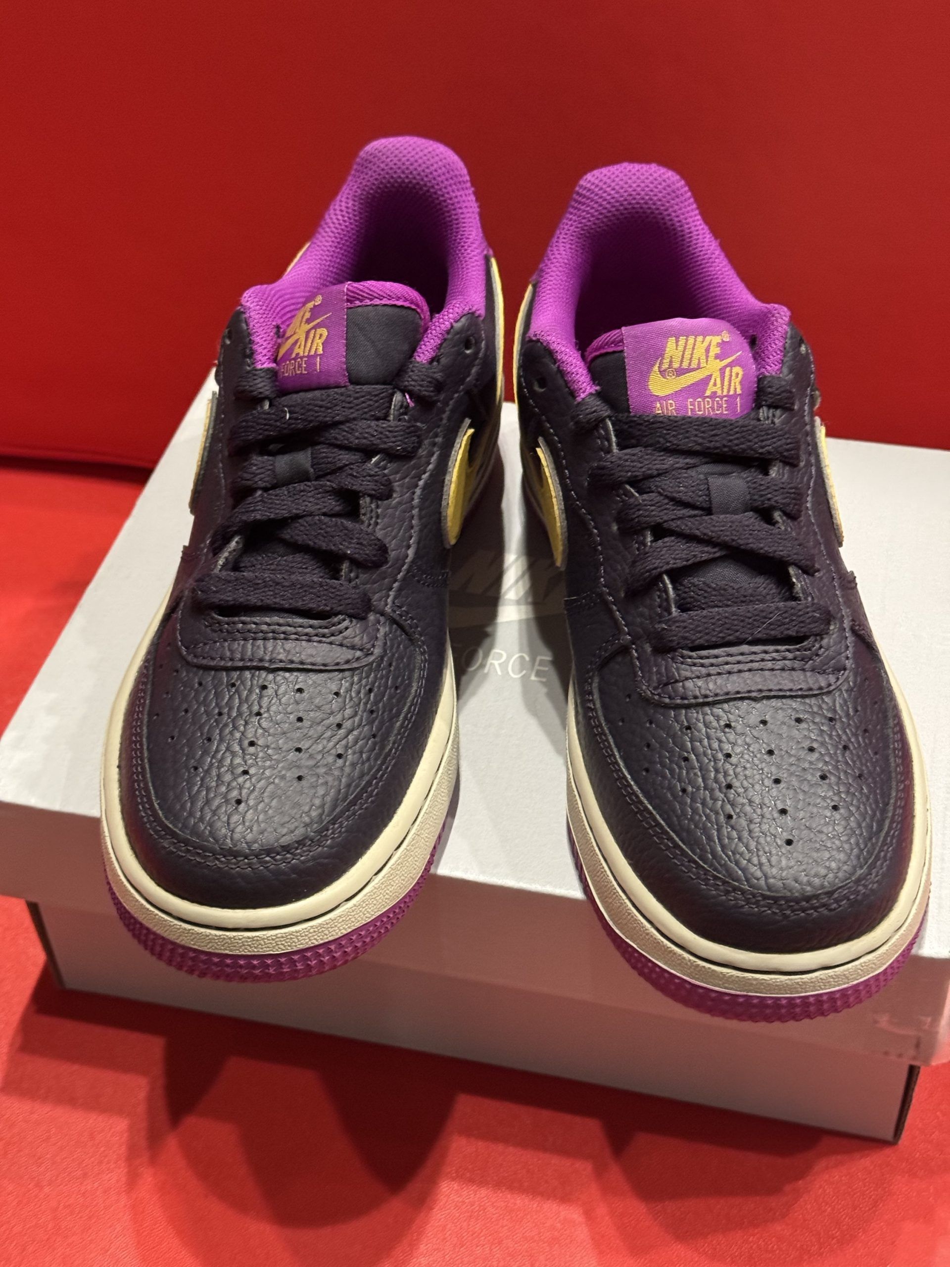 Black and yellow Nike Air Force 1 shoe on a white box, set against a red background. Purple sole.