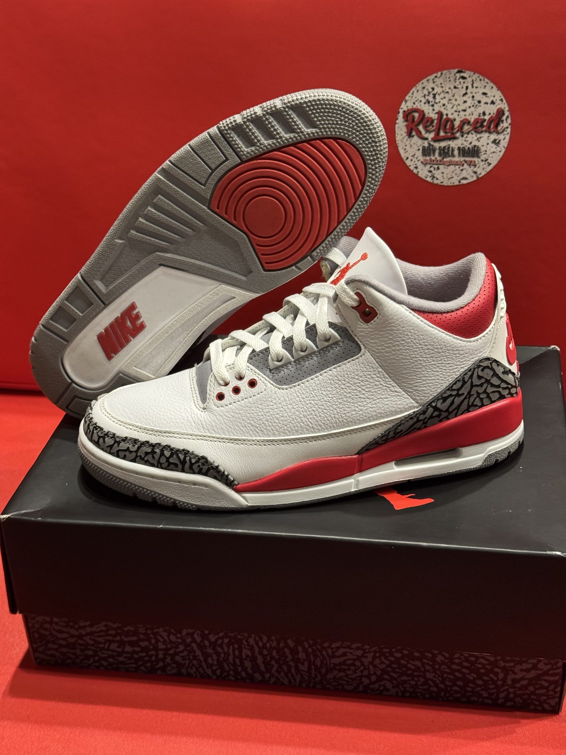 Gray, red, and black Air Jordan 3 sneakers on black box, red background.