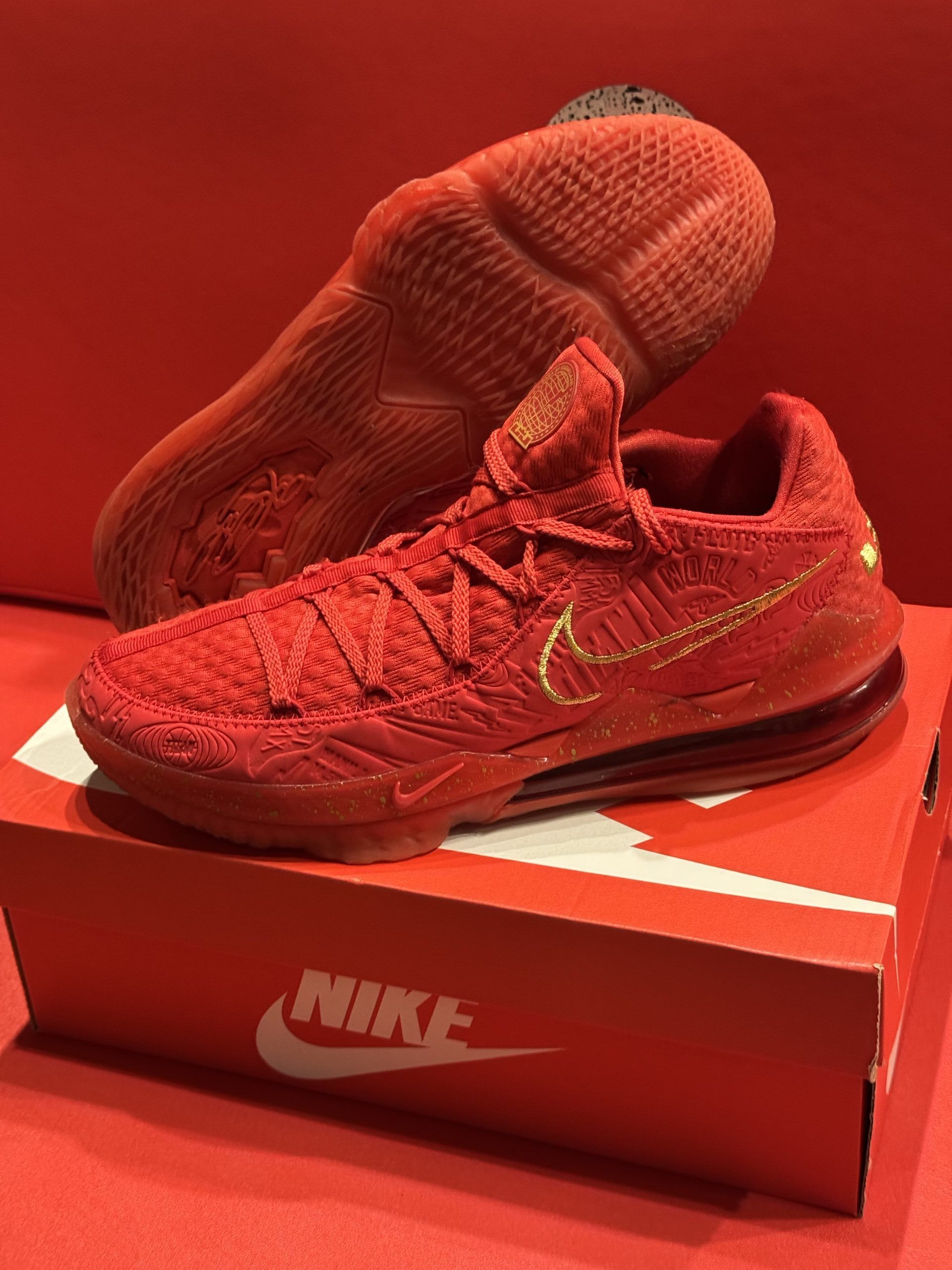 Red Nike LeBron 17 basketball shoe with gold accents on a red box, red background.