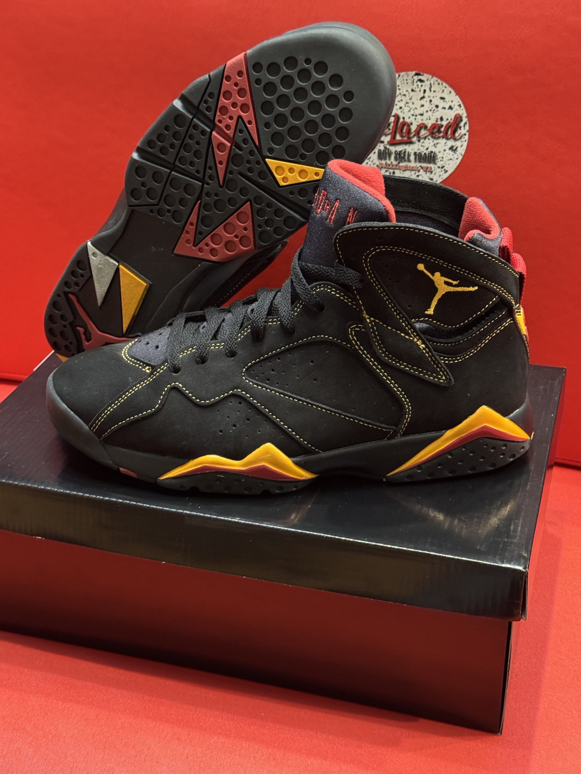 Black and yellow Air Jordan 7 sneakers on a black box, showing the sole. Red background.