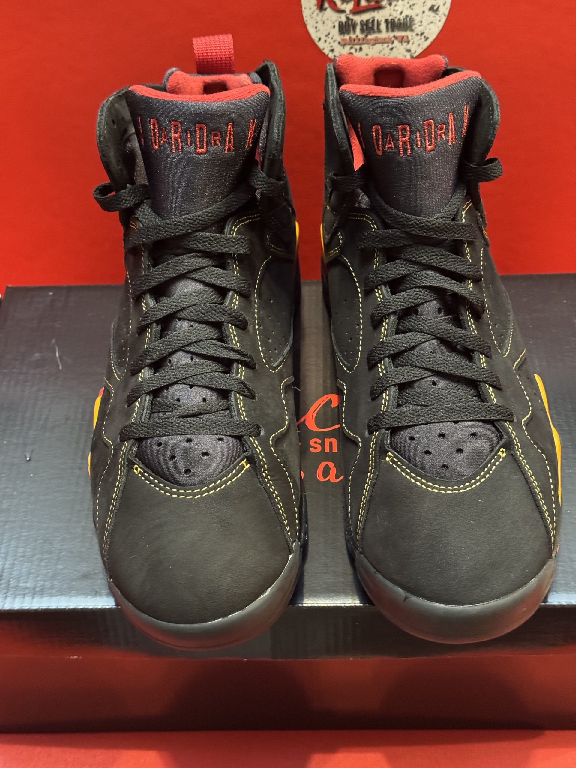 Black and yellow Air Jordan 7 sneakers on a black box, showing the sole. Red background.