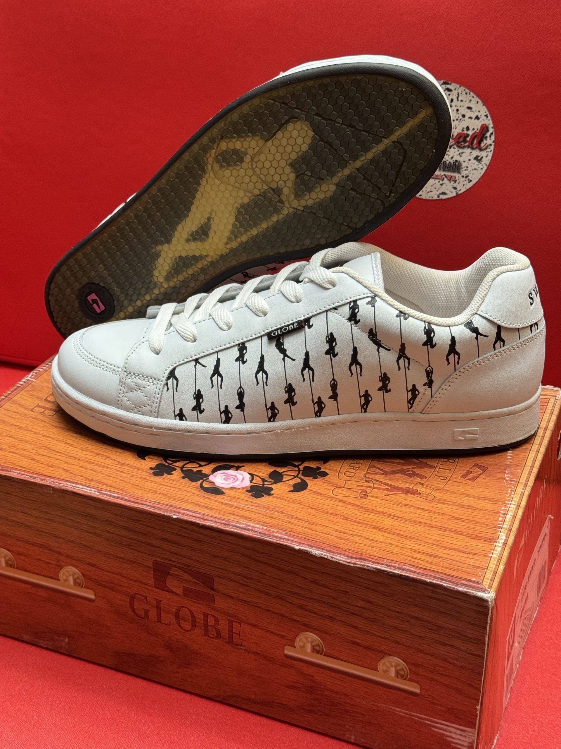 White sneakers with black decorative figures on a wooden box, sole visible, against a red background.