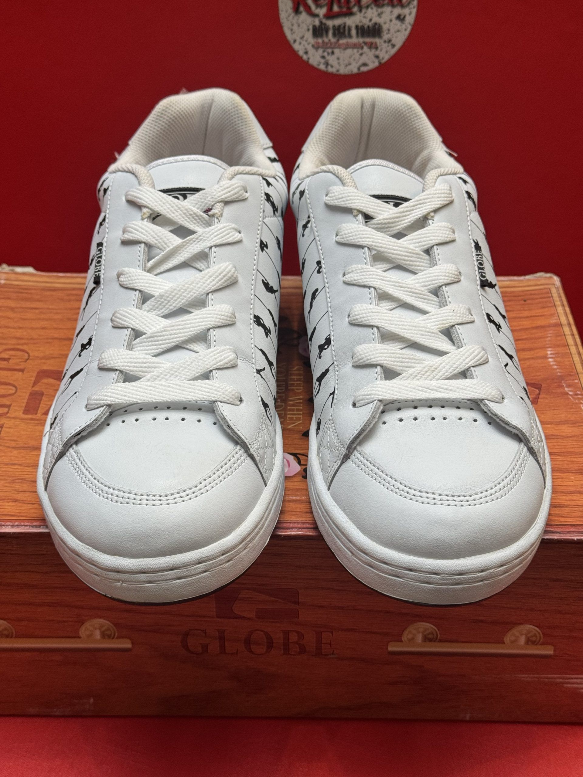 White sneakers with black decorative figures on a wooden box, sole visible, against a red background.