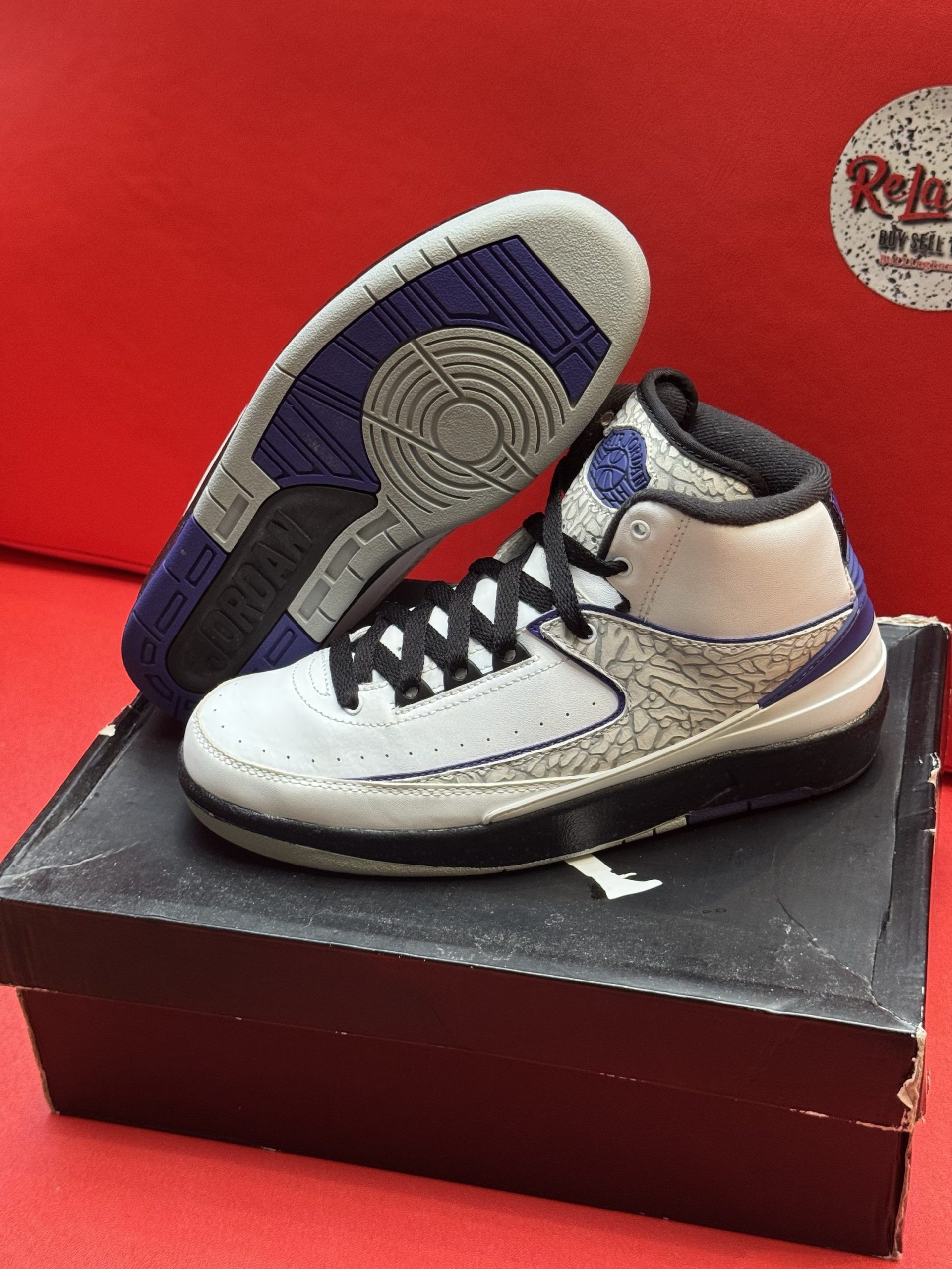 Air Jordan 2 sneakers in white, grey, and purple on a box, sole up.