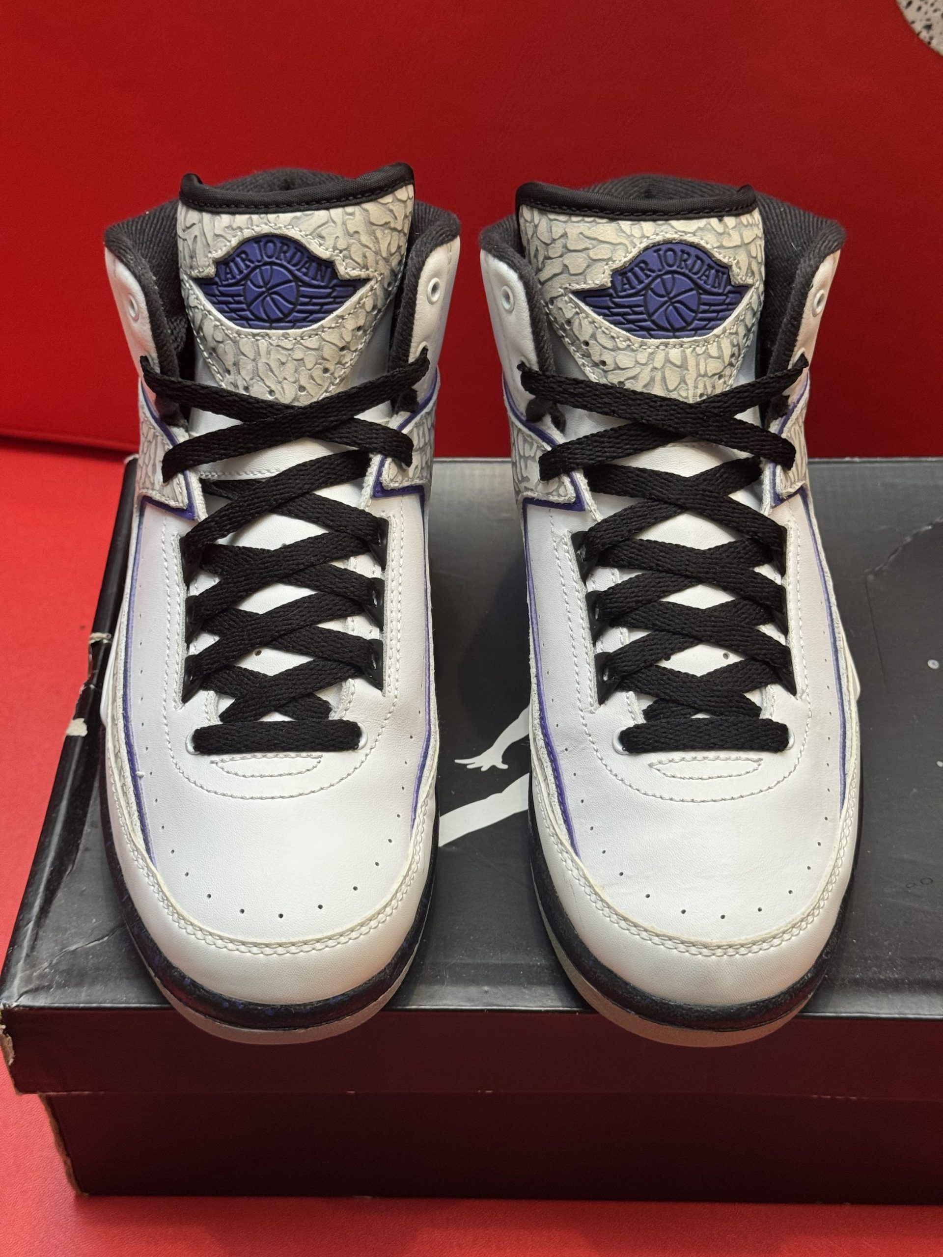 Air Jordan 2 sneakers in white, grey, and purple on a box, sole up.