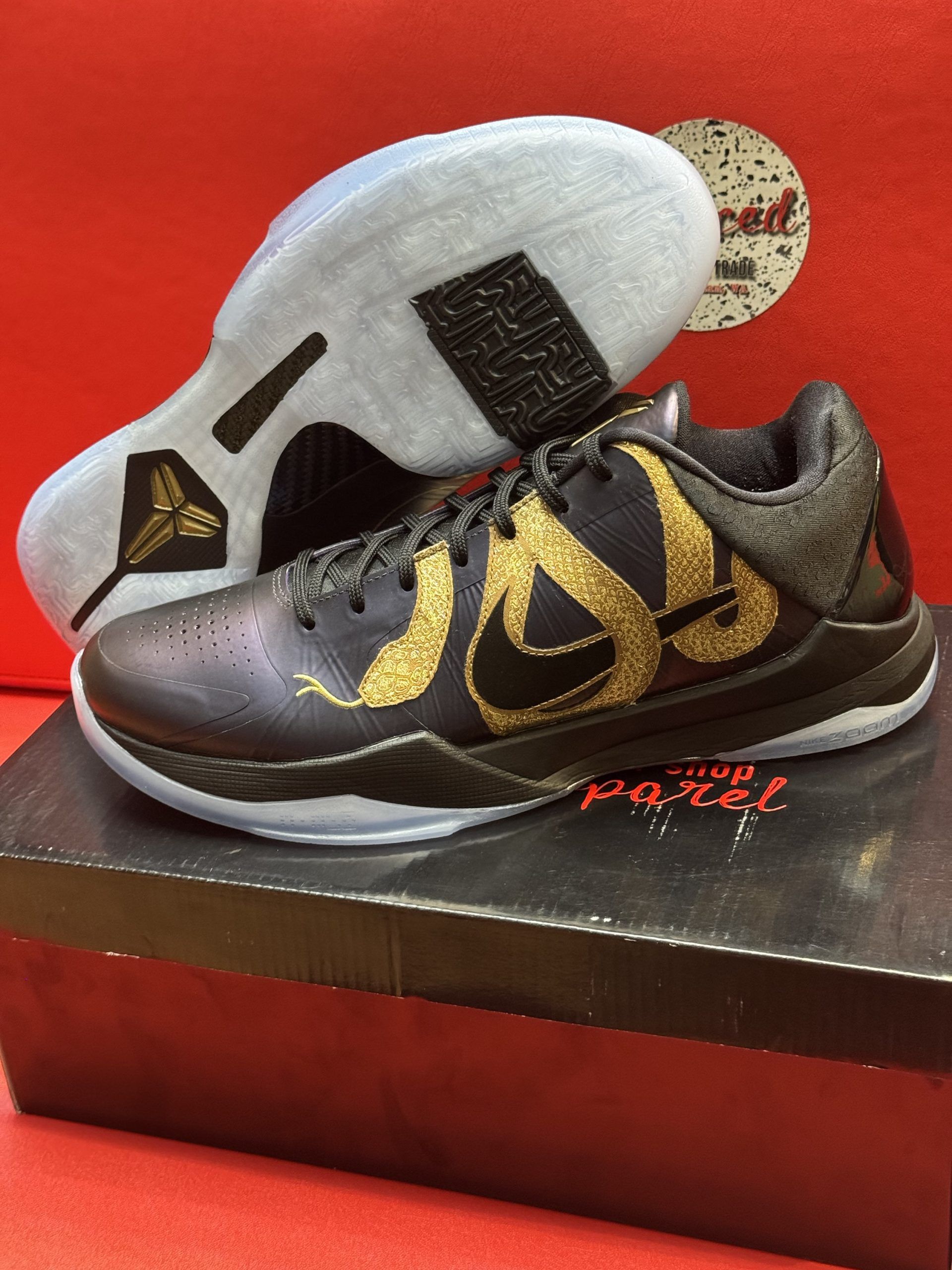 Black and gold Kobe basketball shoes with a translucent sole, displayed on a shoe box against a red backdrop.