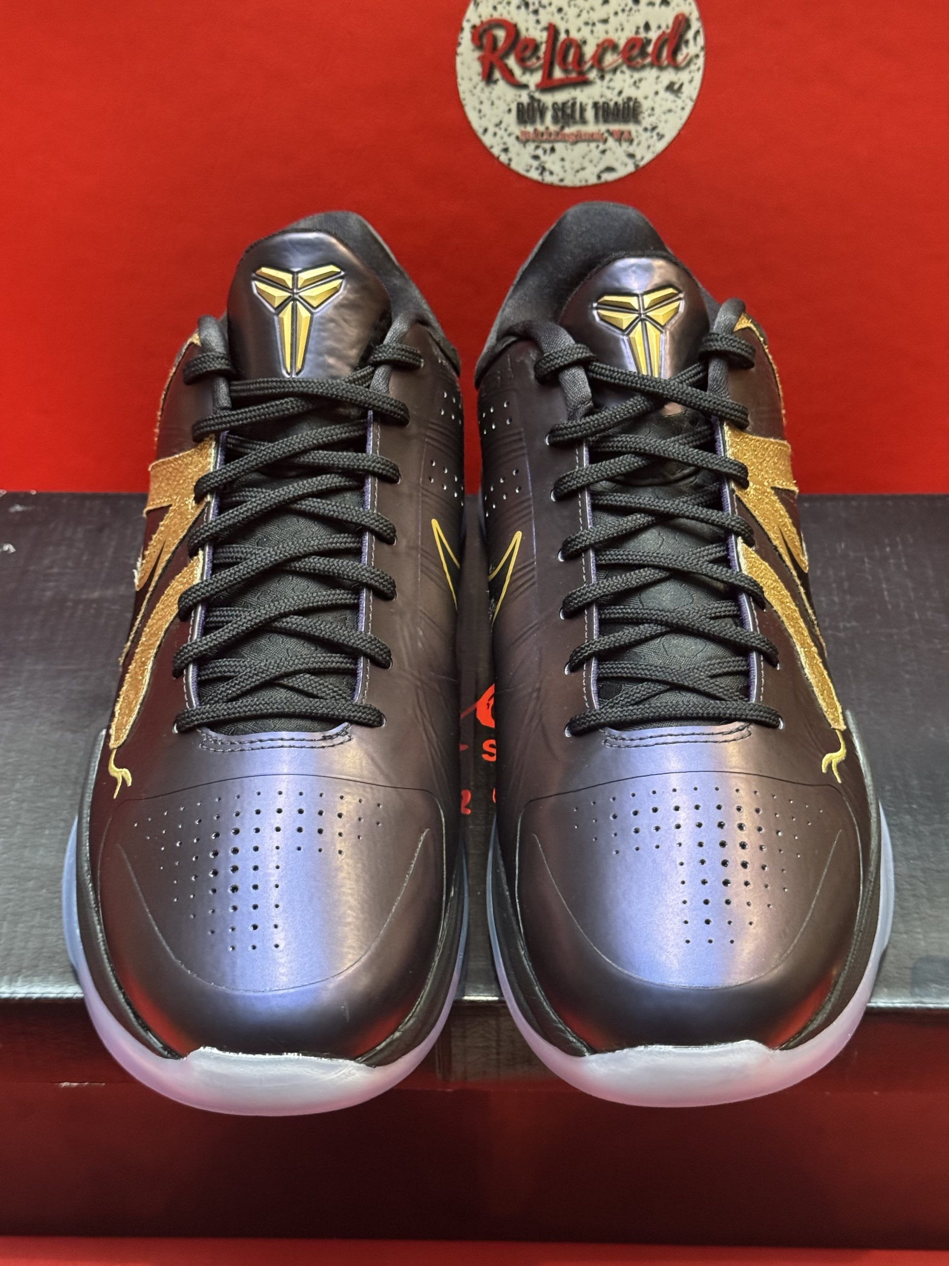 Black and gold Kobe basketball shoes with a translucent sole, displayed on a shoe box against a red backdrop.